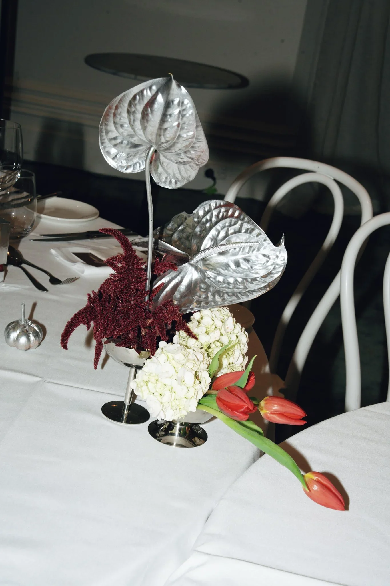 Elegant table centerpiece with silver and red flowers, including red tulips and white hydrangeas, set on a white tablecloth with glassware and cutlery in the background.