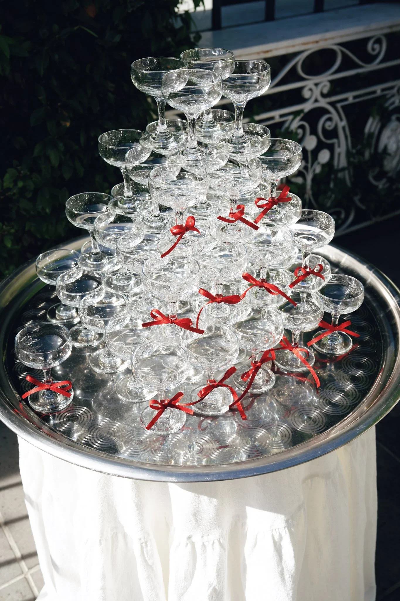 Tiered display of empty champagne glasses decorated with red ribbons on a silver tray with a white cloth underneath, set outdoors at daytime.
