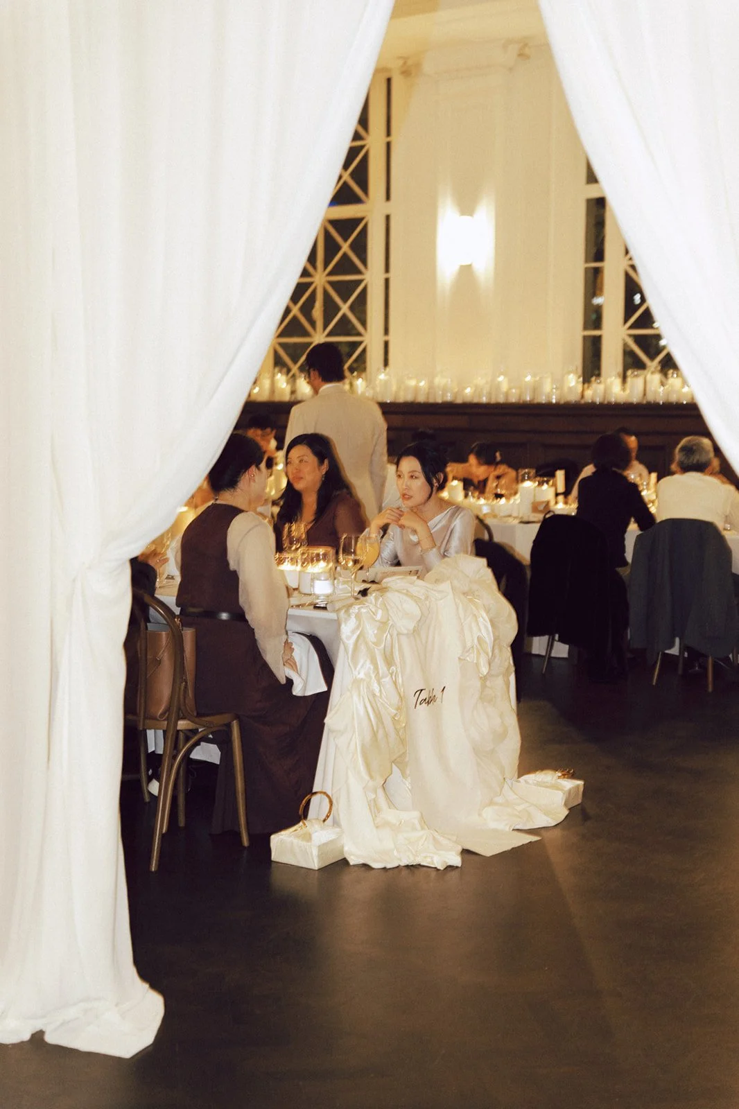 People gathered at a formal dinner event, seen through white drapes, with candles on tables and a large window in the background.
