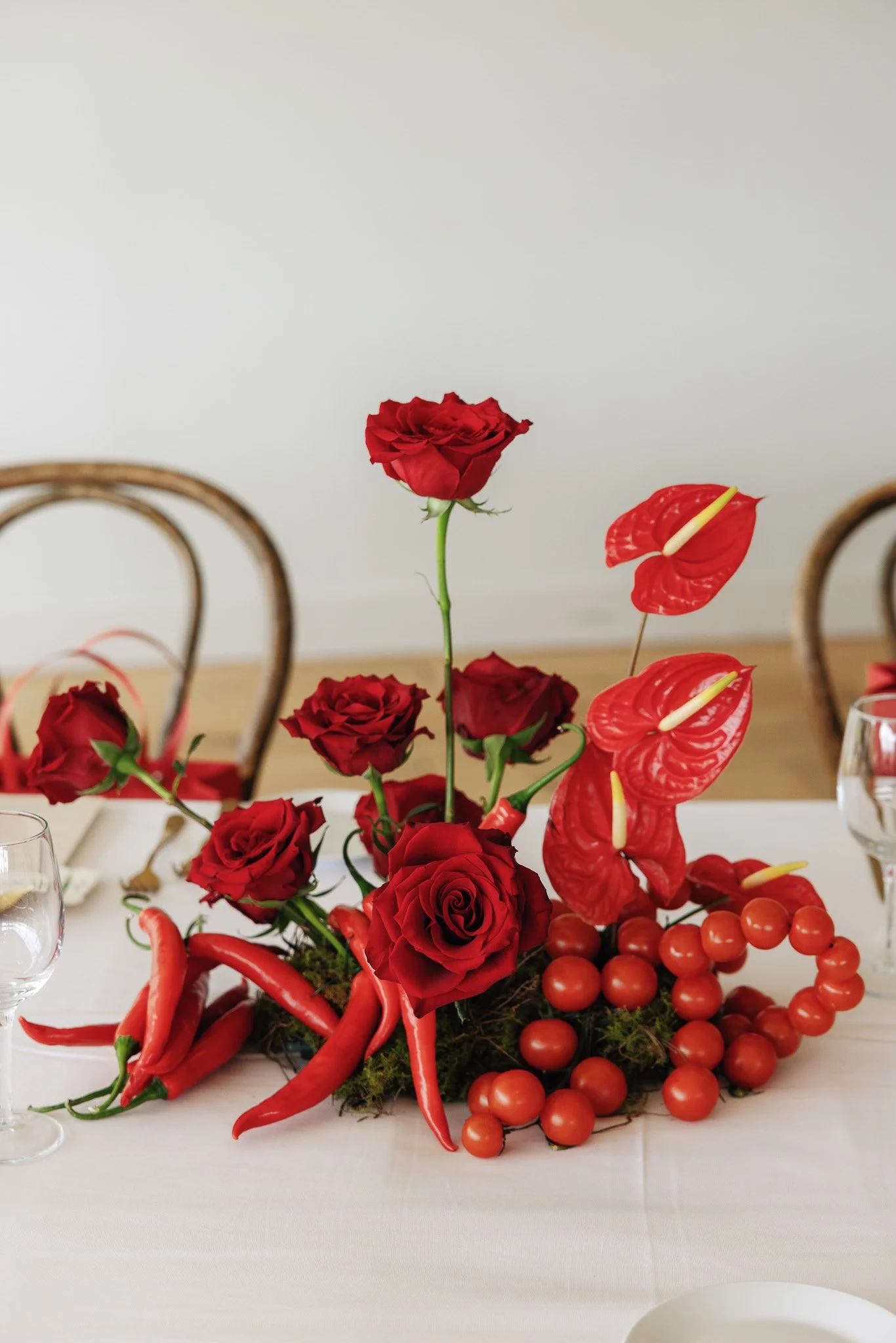A table centerpiece with red roses, red anthuriums, orange cherry tomatoes, and red chili peppers arranged on a bed of moss, with chairs and wine glasses visible in the background.