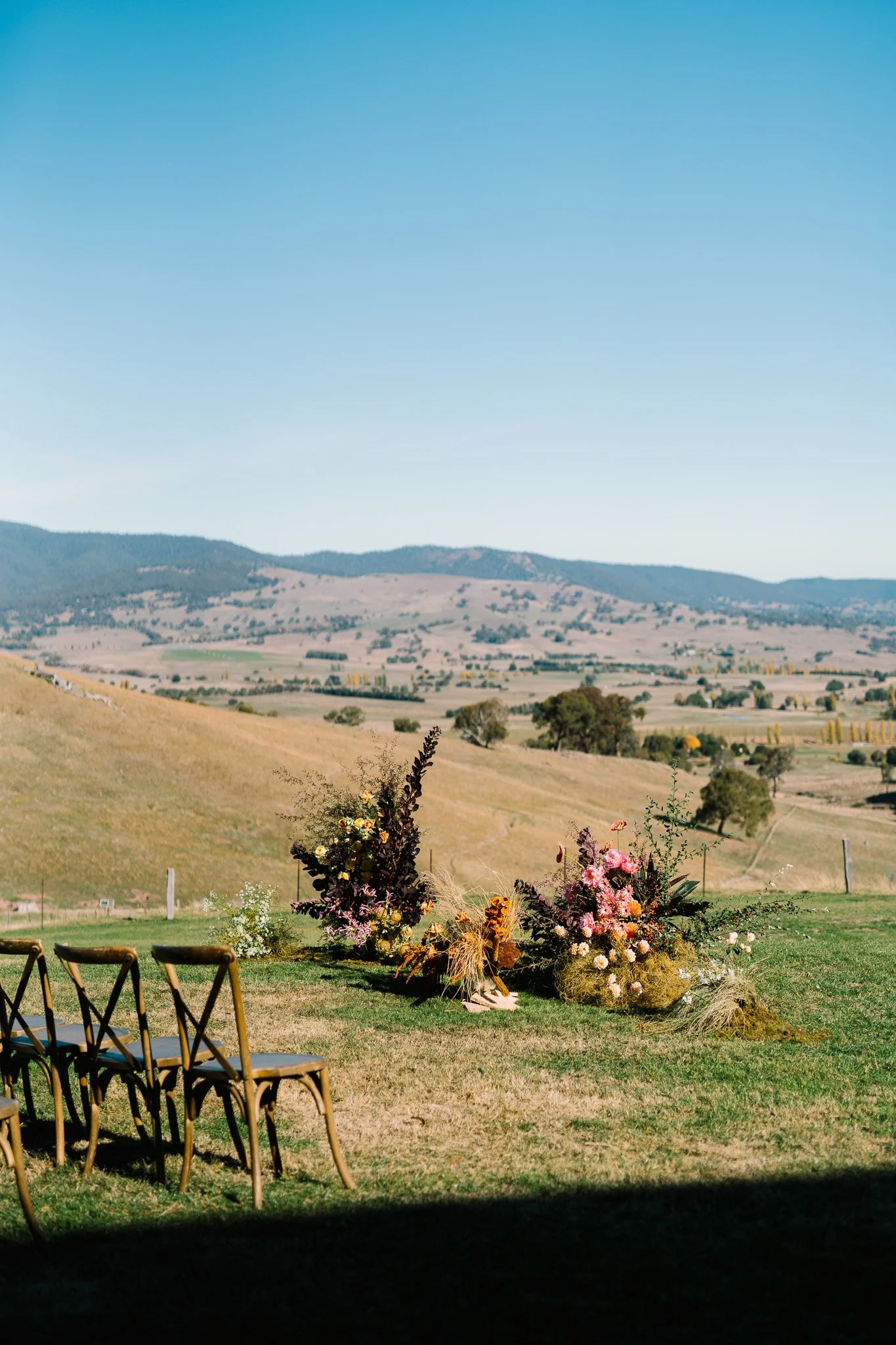 A scenic outdoor landscape with a distant view of rolling hills under a blue sky. In the foreground, there is a floral arrangement with various flowers and foliage, and a few chairs are set up on the grass, suggesting a setup for an outdoor event or 