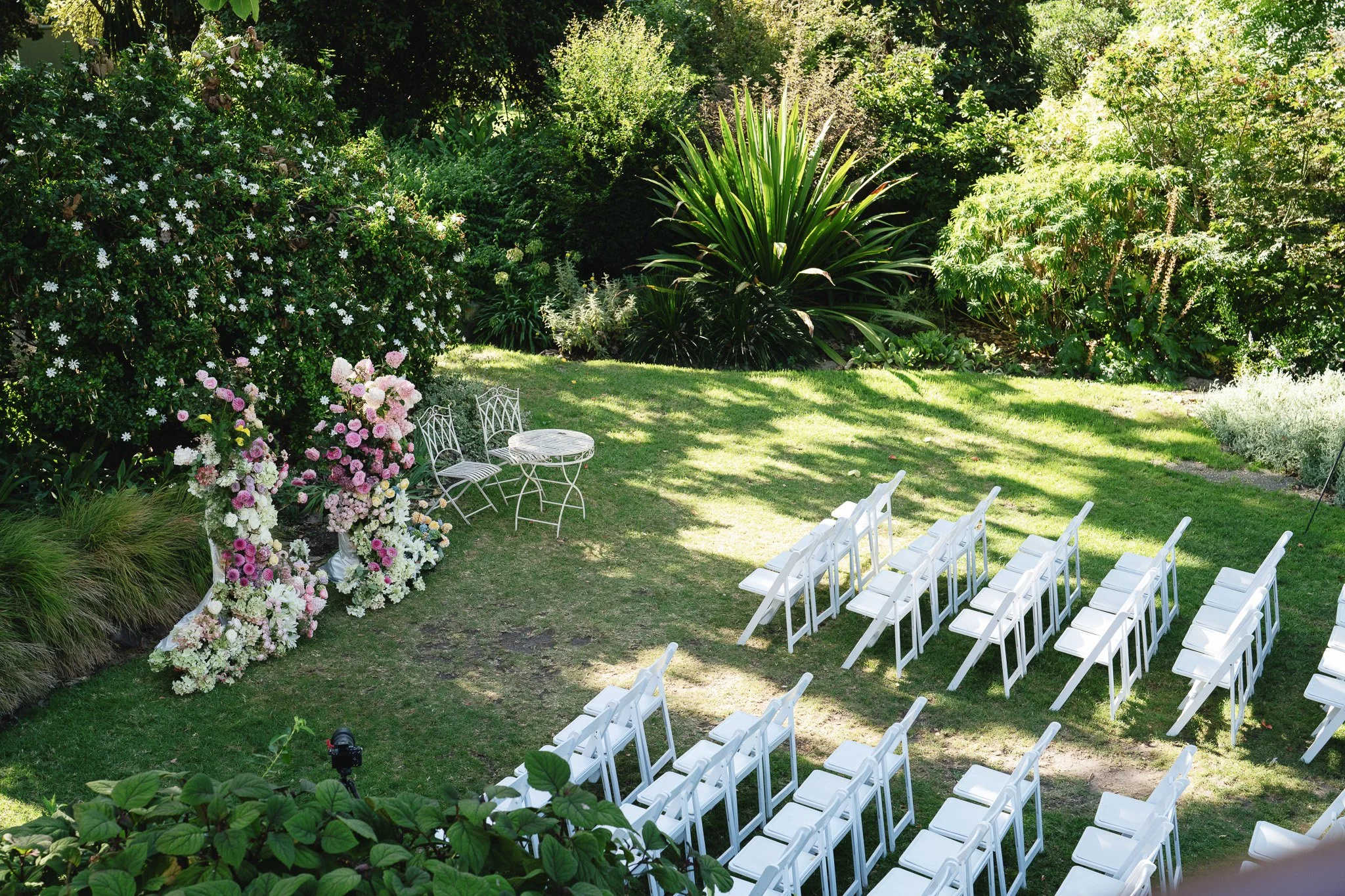 A garden setup with white chairs arranged in rows on the grass, floral arrangements on the left side, and lush green bushes and trees in the background, indicating an outdoor event space.