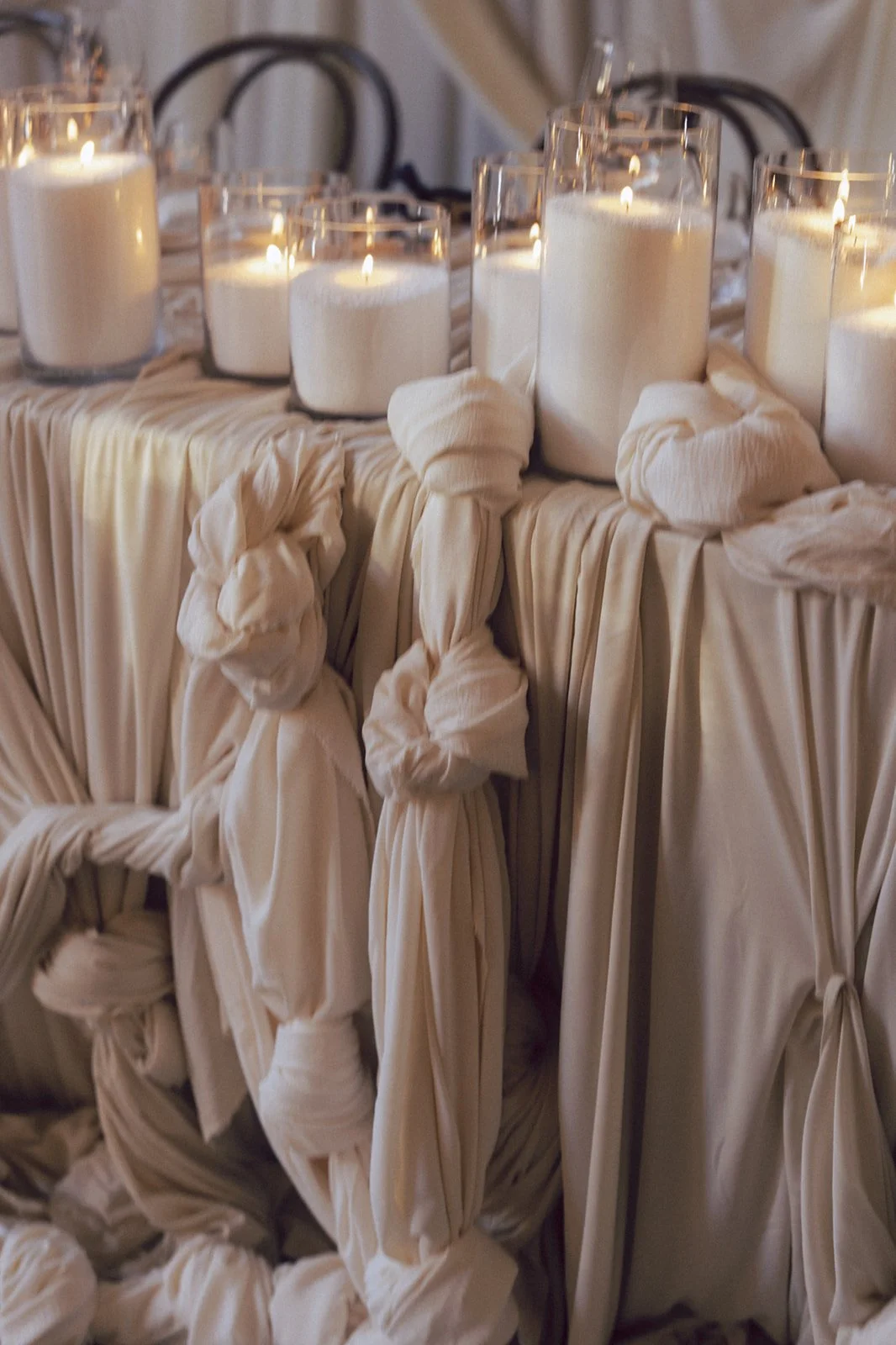 Decorative table with white candles in glass holders, set on a draped ivory fabric with knotted and pleated details.