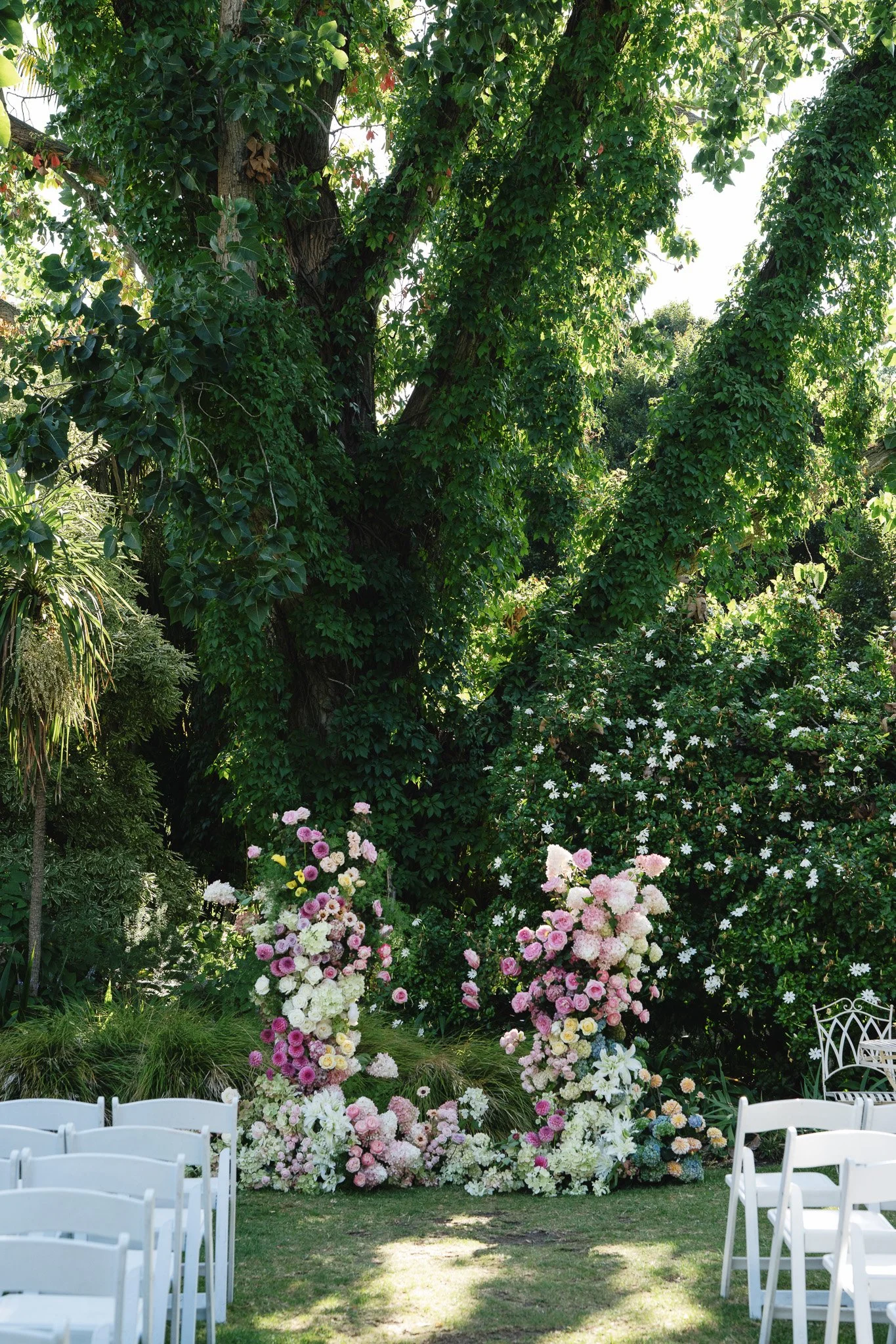 Outdoor wedding setup with white chairs arranged in rows facing a floral arch and lush green trees with sunlight filtering through.