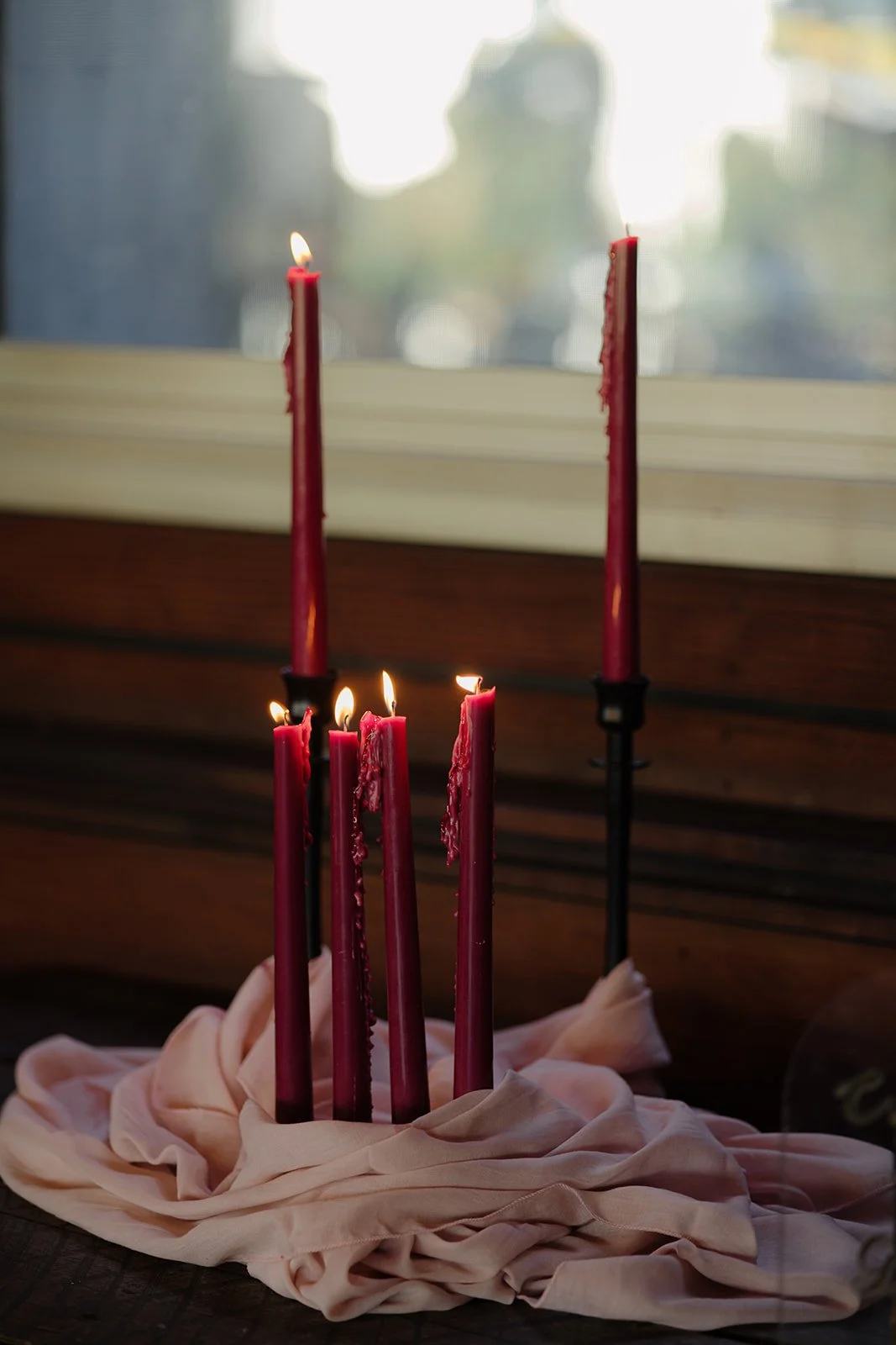 Pink birthday candles, some lit and some melted down, are placed on a cloth on a wooden surface in front of a window with blurred outdoor scenery.