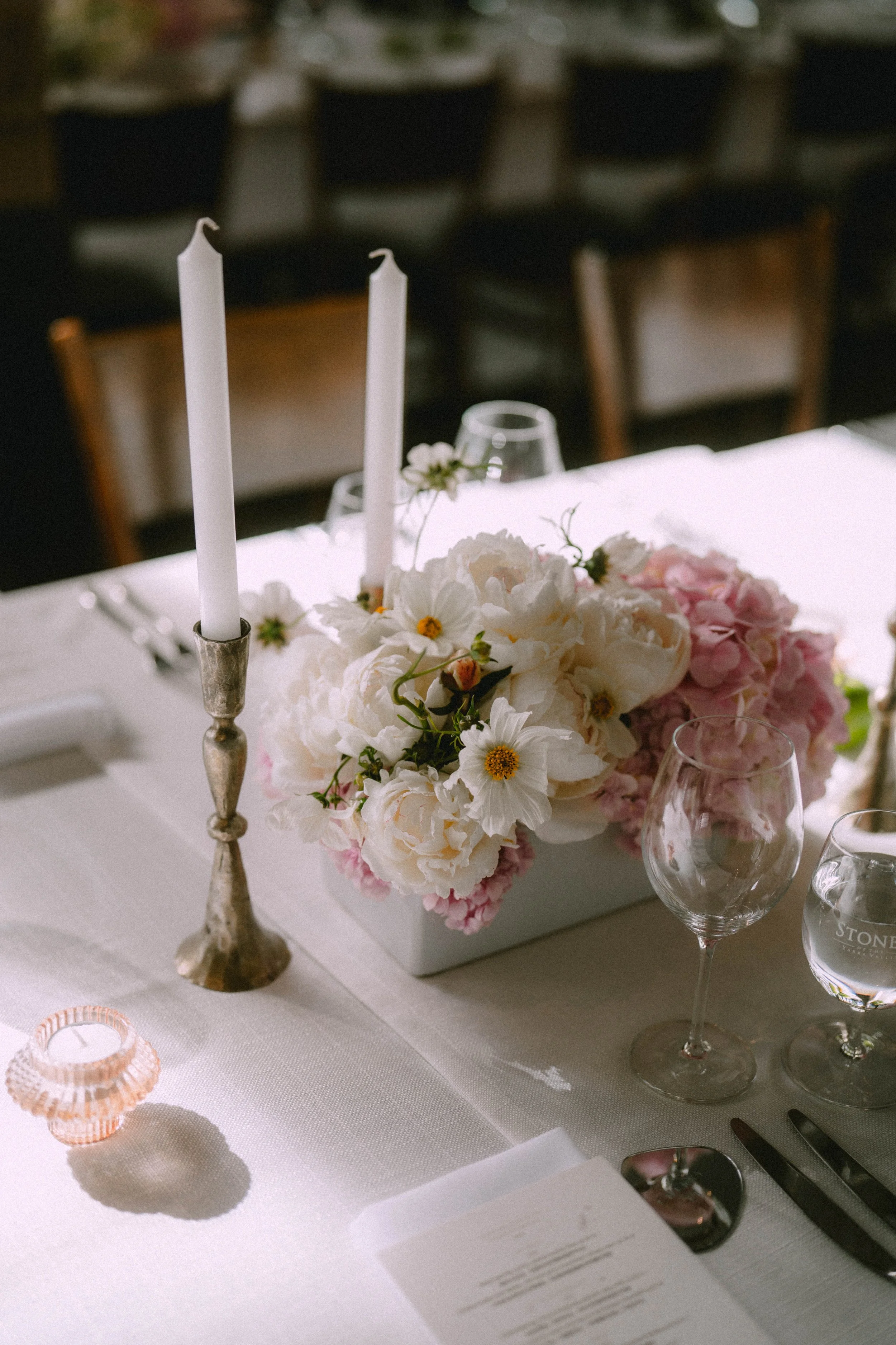 A table decorated with a floral centerpiece consisting of white and pink flowers, a silver candlestick with white candles, wine glasses, and a menu in a well-lit setting for a special occasion or celebration.