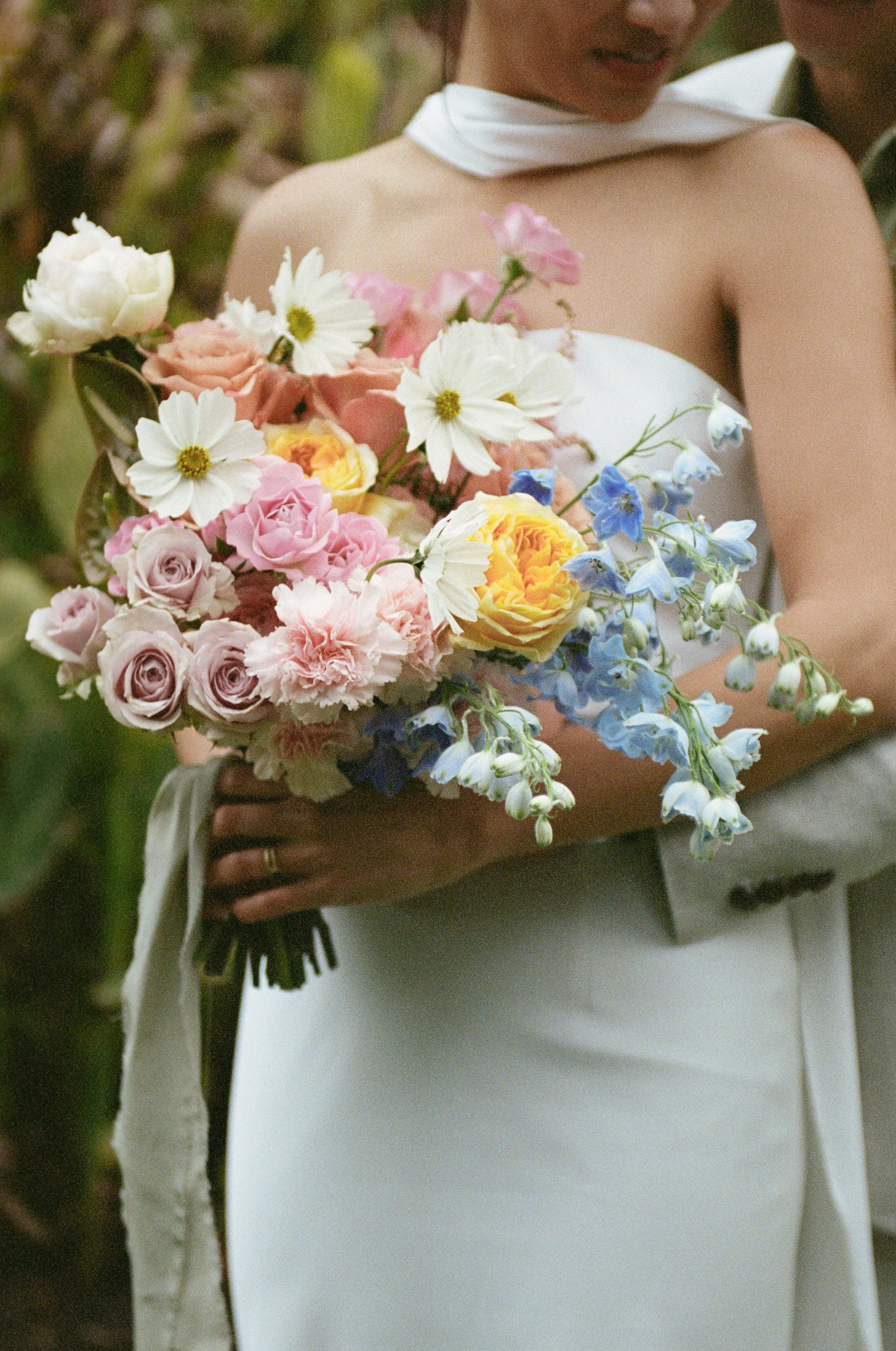 A woman wearing a white dress and a white scarf around her neck is holding a bouquet of mixed flowers including white, pink, yellow roses, and blue delphiniums. She is outdoors, with greenery in the background.