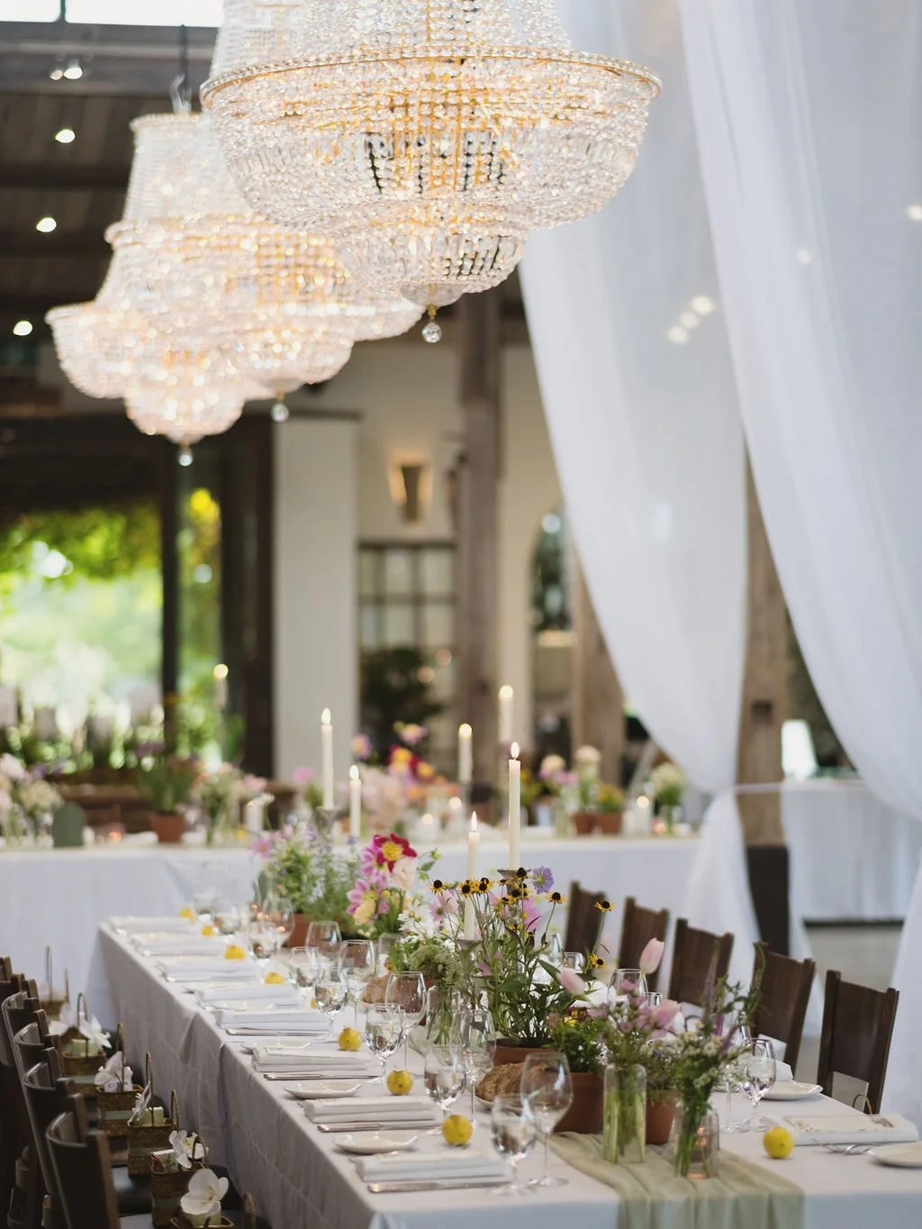 Elegant banquet table decorated with fresh flowers, candles, and glassware under large chandeliers in a bright, airy venue.