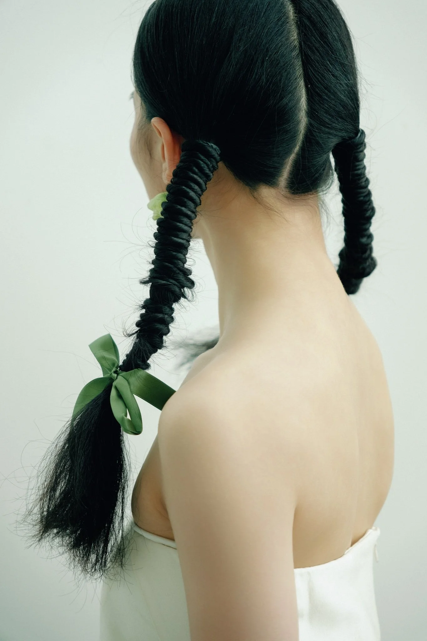 Back of a mannequin with long black braided hair tied with a green ribbon, wearing a white strapless top.