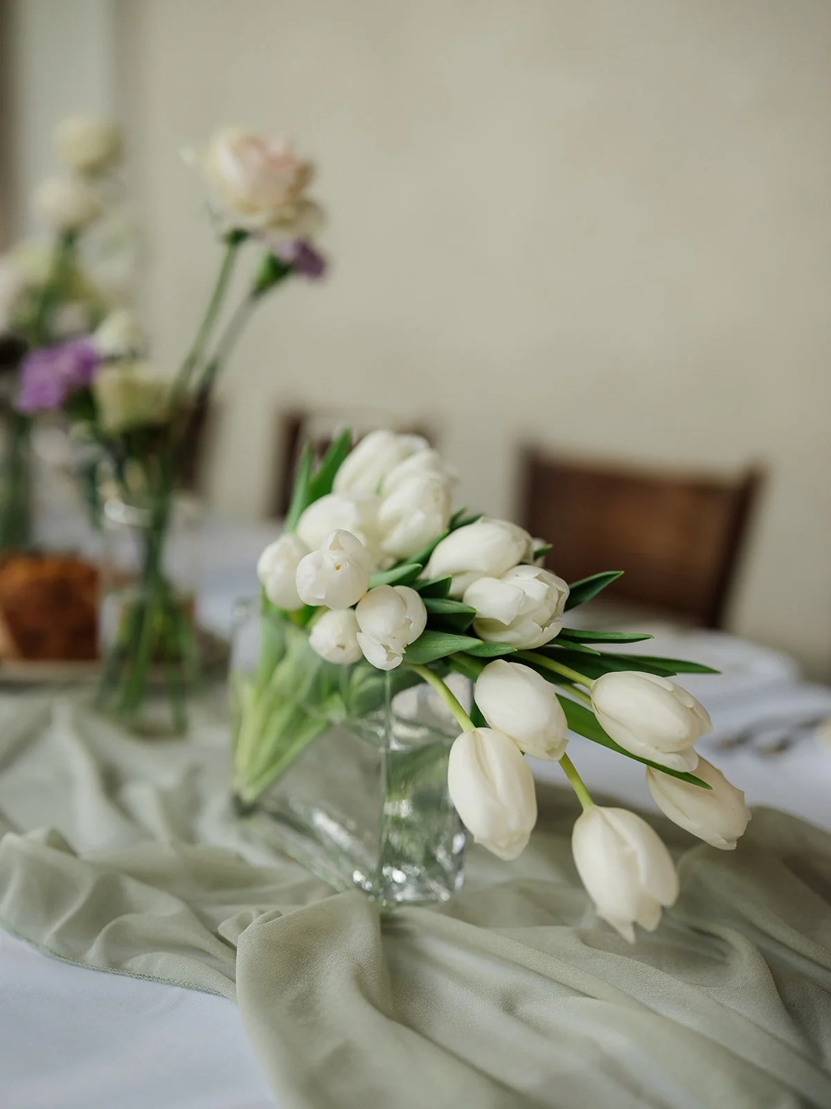 White tulip bouquet in a clear glass pitcher on a decorated table.