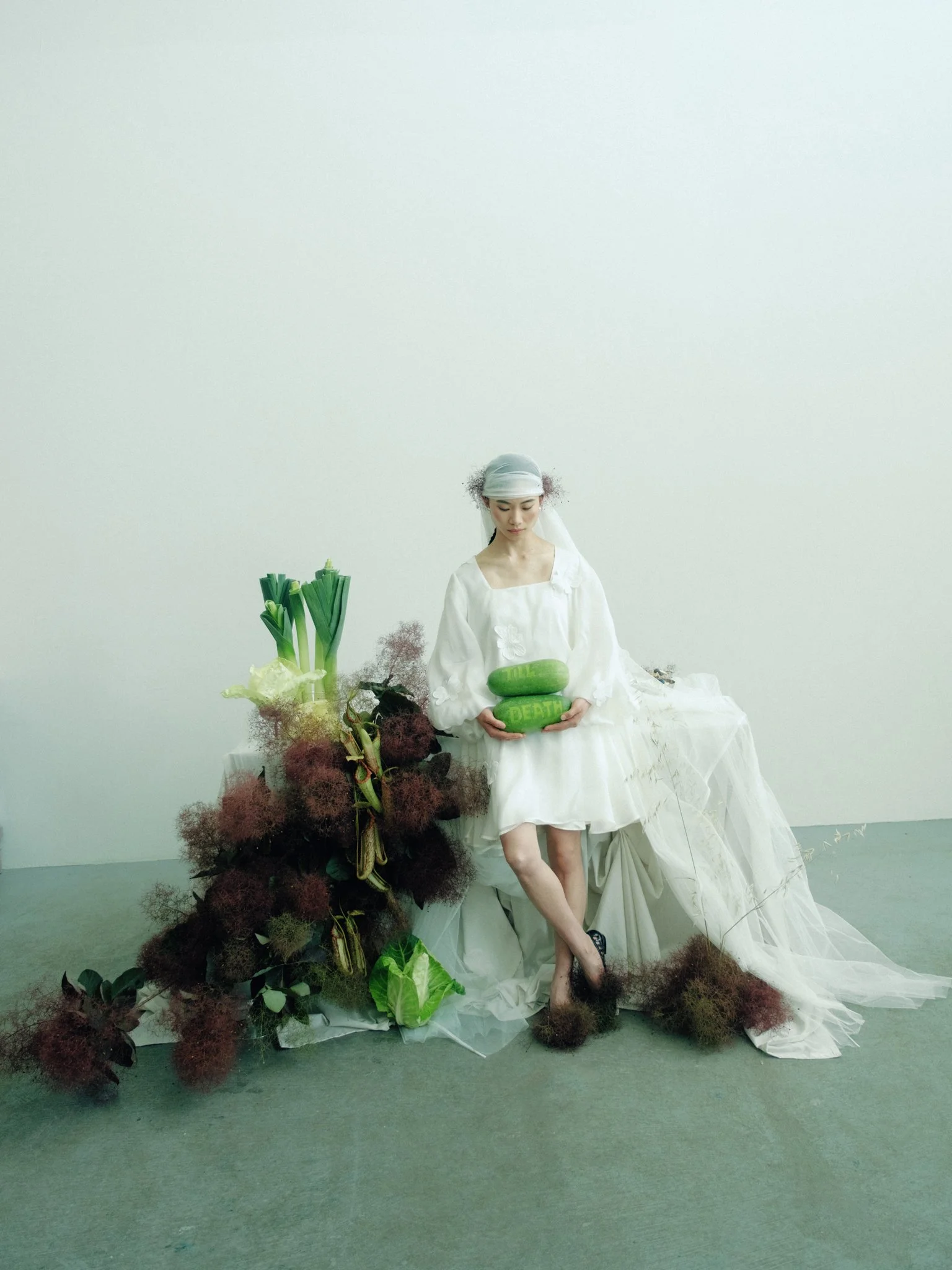A woman dressed in white, sitting amongst large green vegetables, including leeks and lettuce, with dark red and brown plant like structures around her, in a minimalist white room.
