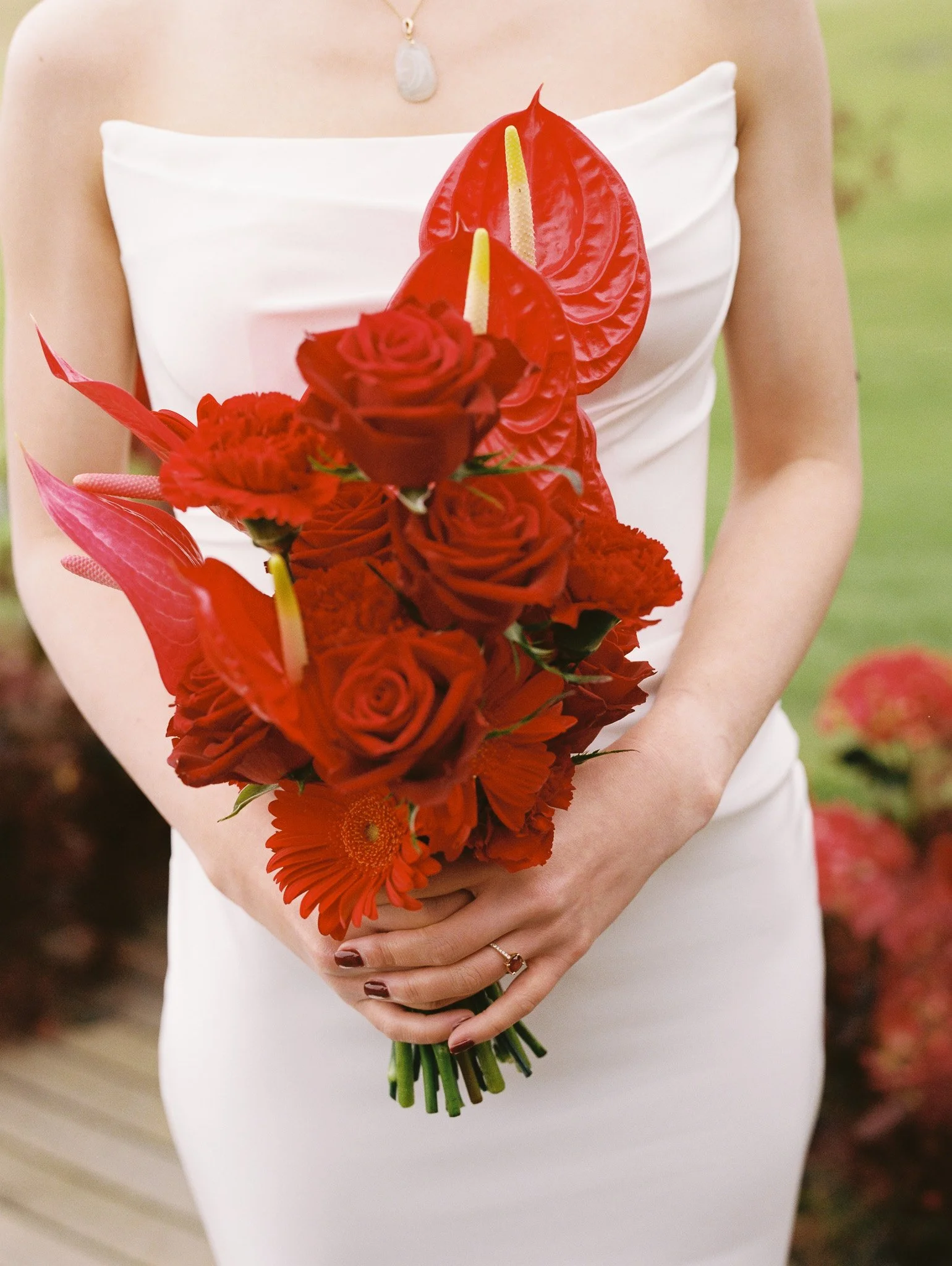 Person in a white dress holding a bouquet of red flowers, including roses and anthuriums, outdoors.