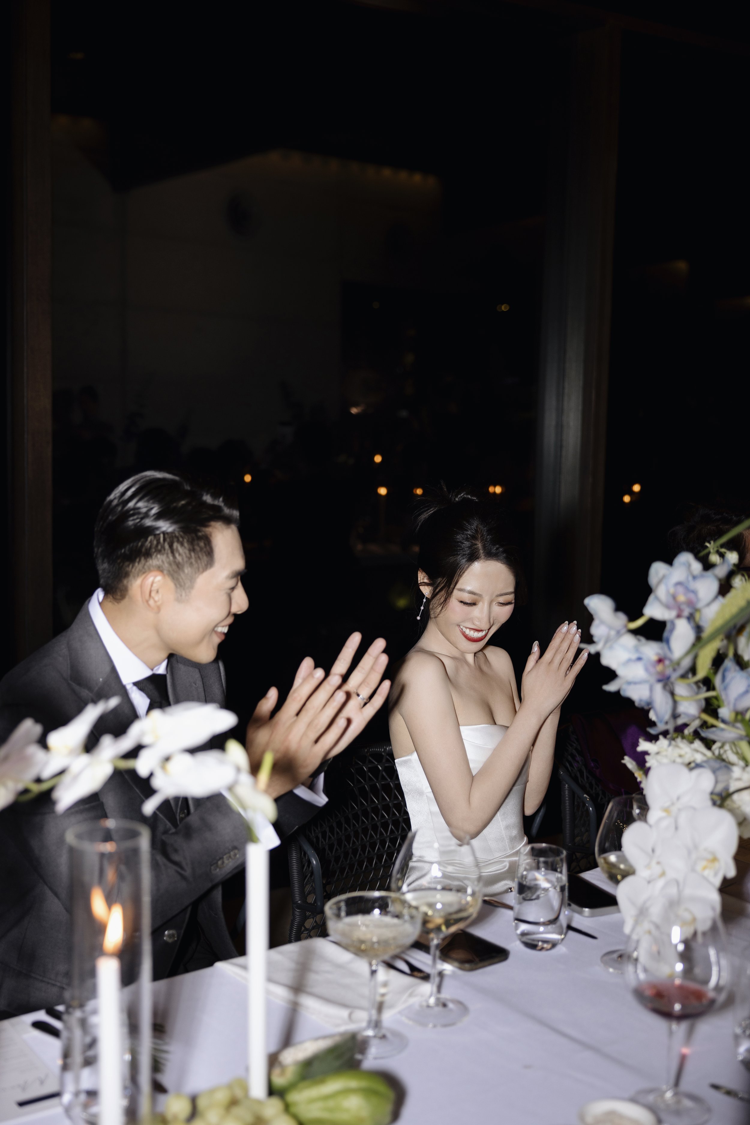 A group of people dressed in formal attire sitting at a dining table during a celebration or event, possibly a wedding reception. The table has white tablecloths, flower arrangements, candles, and glasses of wine.