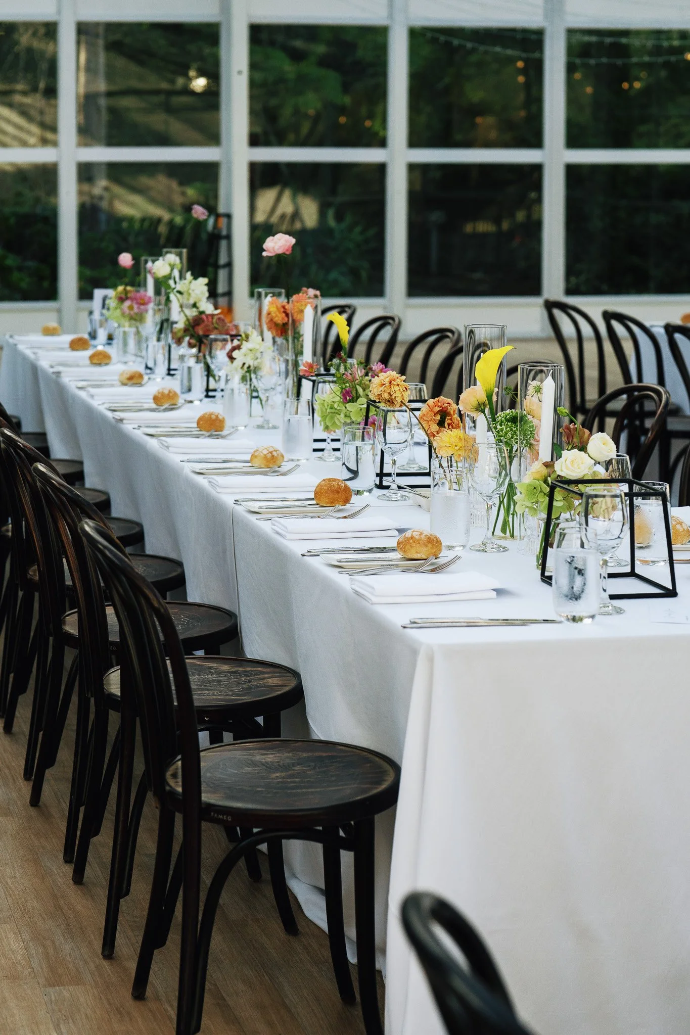 Long banquet table with white tablecloth, decorated with colorful flowers in vases, and set with bread rolls, glasses, and silverware, in a bright room with large windows and black chairs.