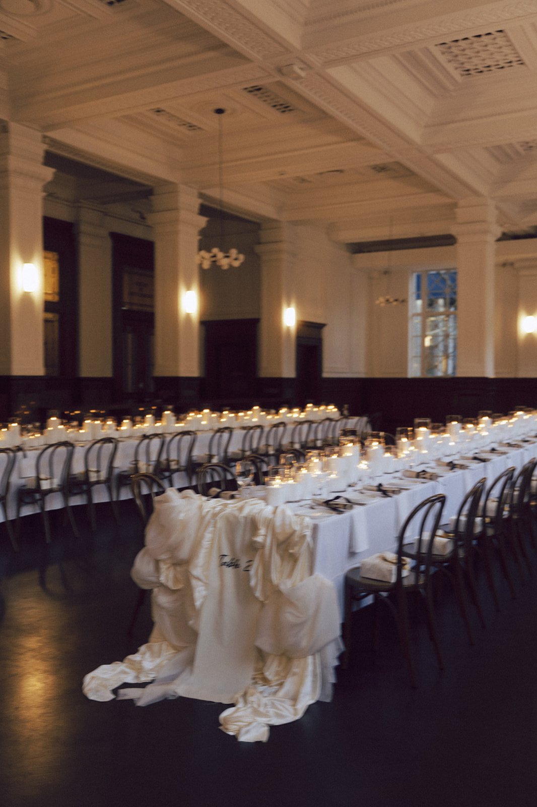 A long banquet table set for a formal event with white tablecloths, candles, and place settings in a decorated hall with high ceiling and lighting fixtures.