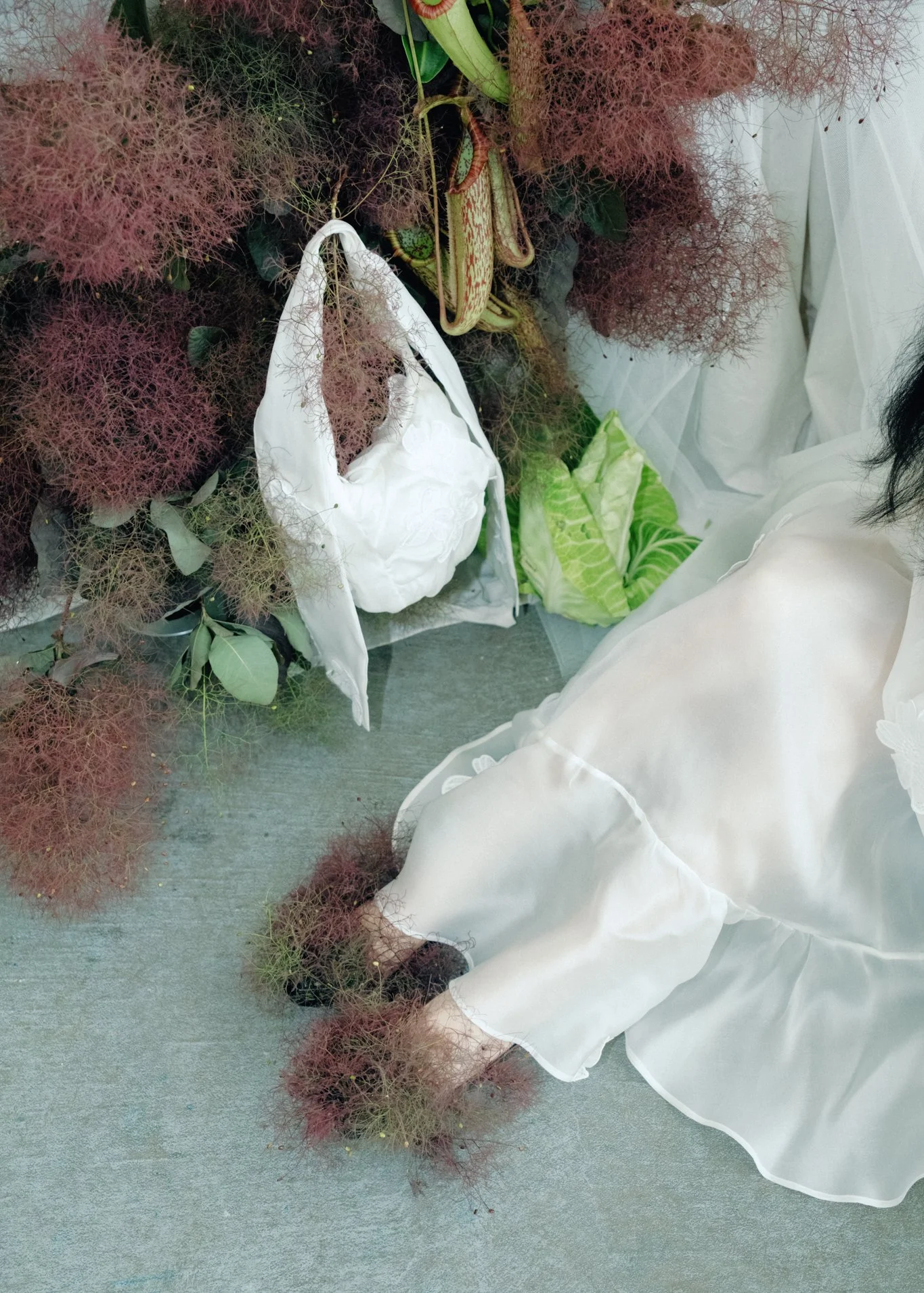 A person wearing a white dress and black hair, is arranging a floral display that includes pink, delicate, wispy foliage, a white fabric bag, and green leaves, in a space with a gray floor and white backdrop.