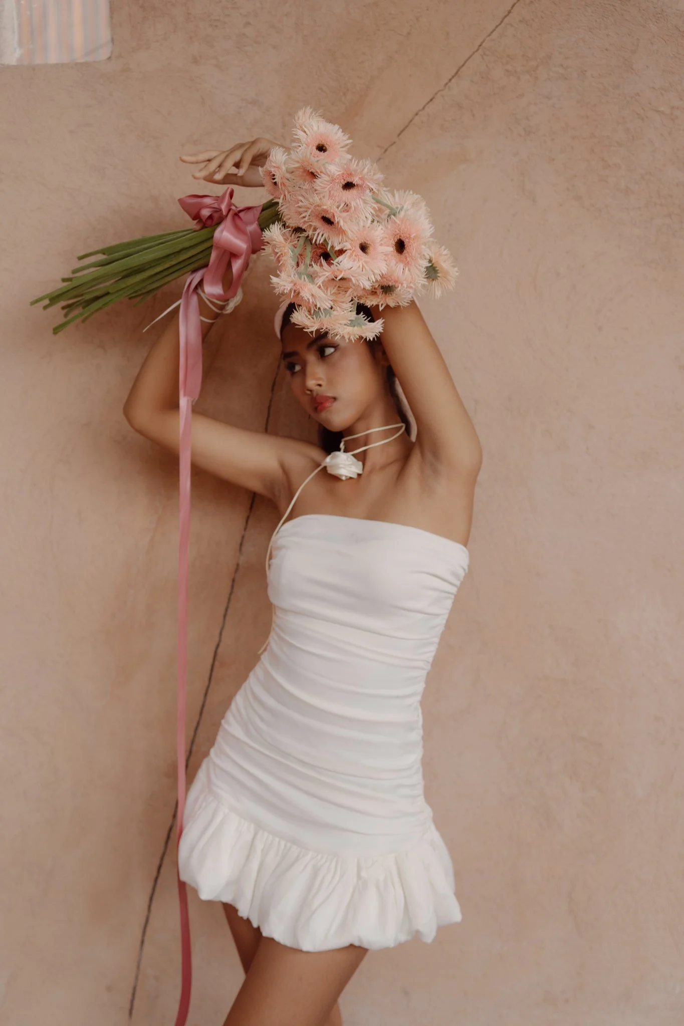 A woman in a white strapless dress holding a large bouquet of pink flowers with long green stems and pink ribbon, standing against a beige wall.