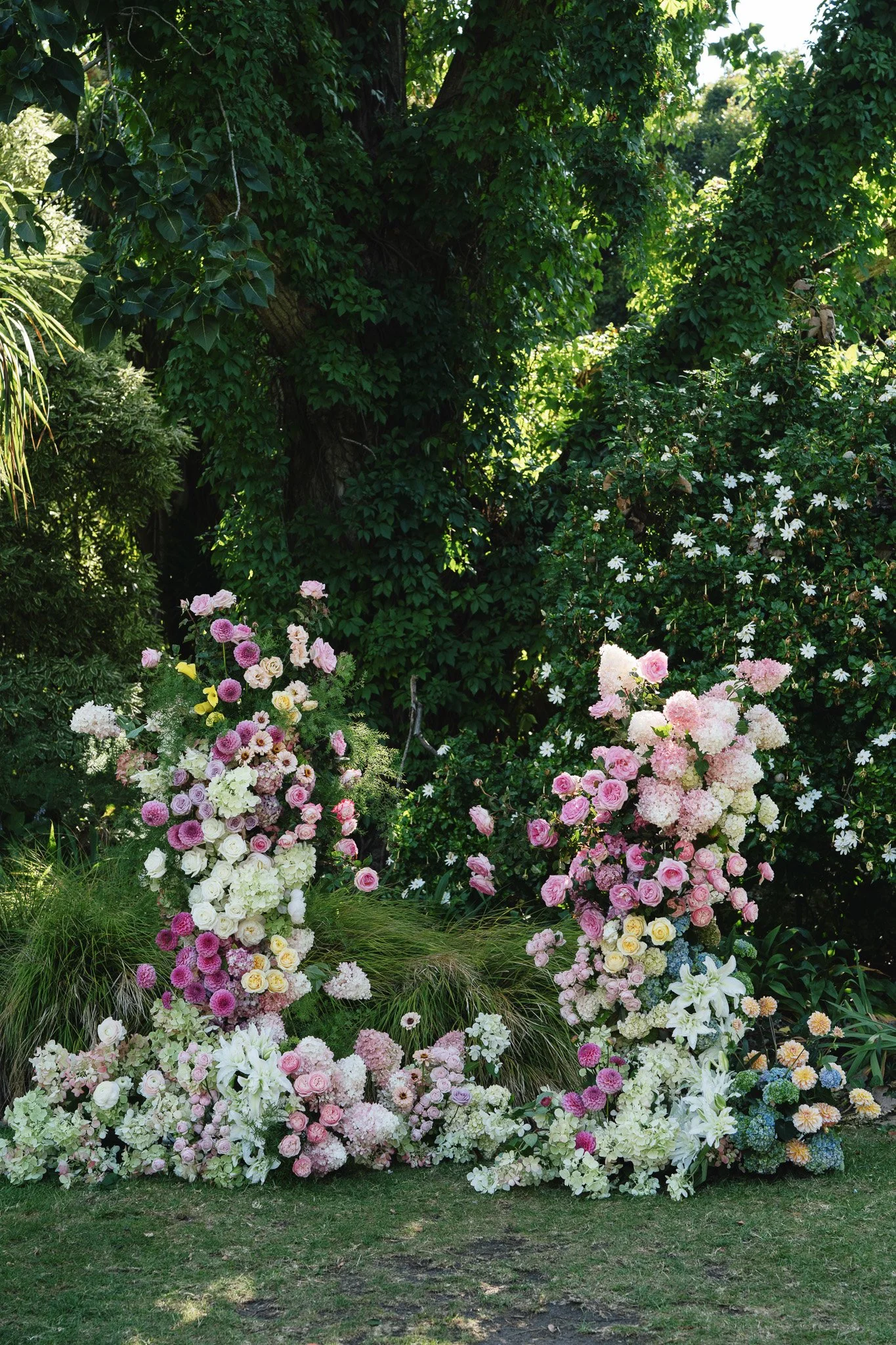 A garden with a large, leafy tree in the background and numerous colorful flowers, including pink and white roses and hydrangeas, arranged in natural flower beds.
