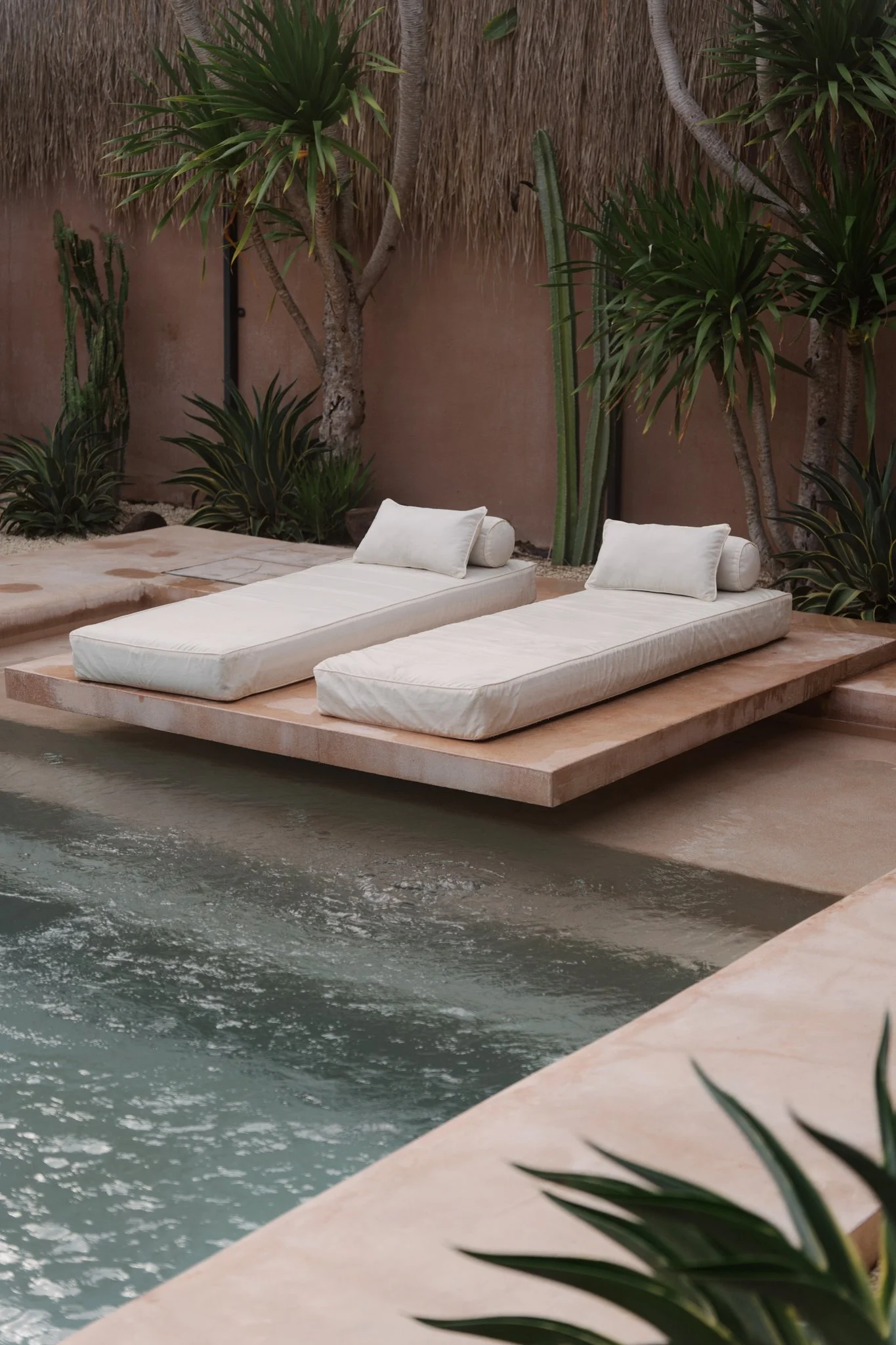 Two white lounge chairs with pillows next to a pool, surrounded by tropical plants and trees.
