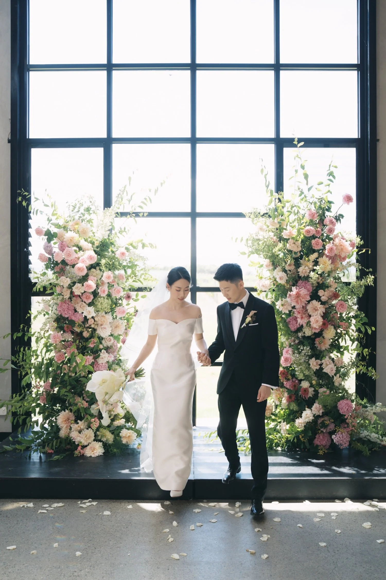 A bride and groom standing hand in hand in front of a large window adorned with floral arrangements, during a wedding ceremony.