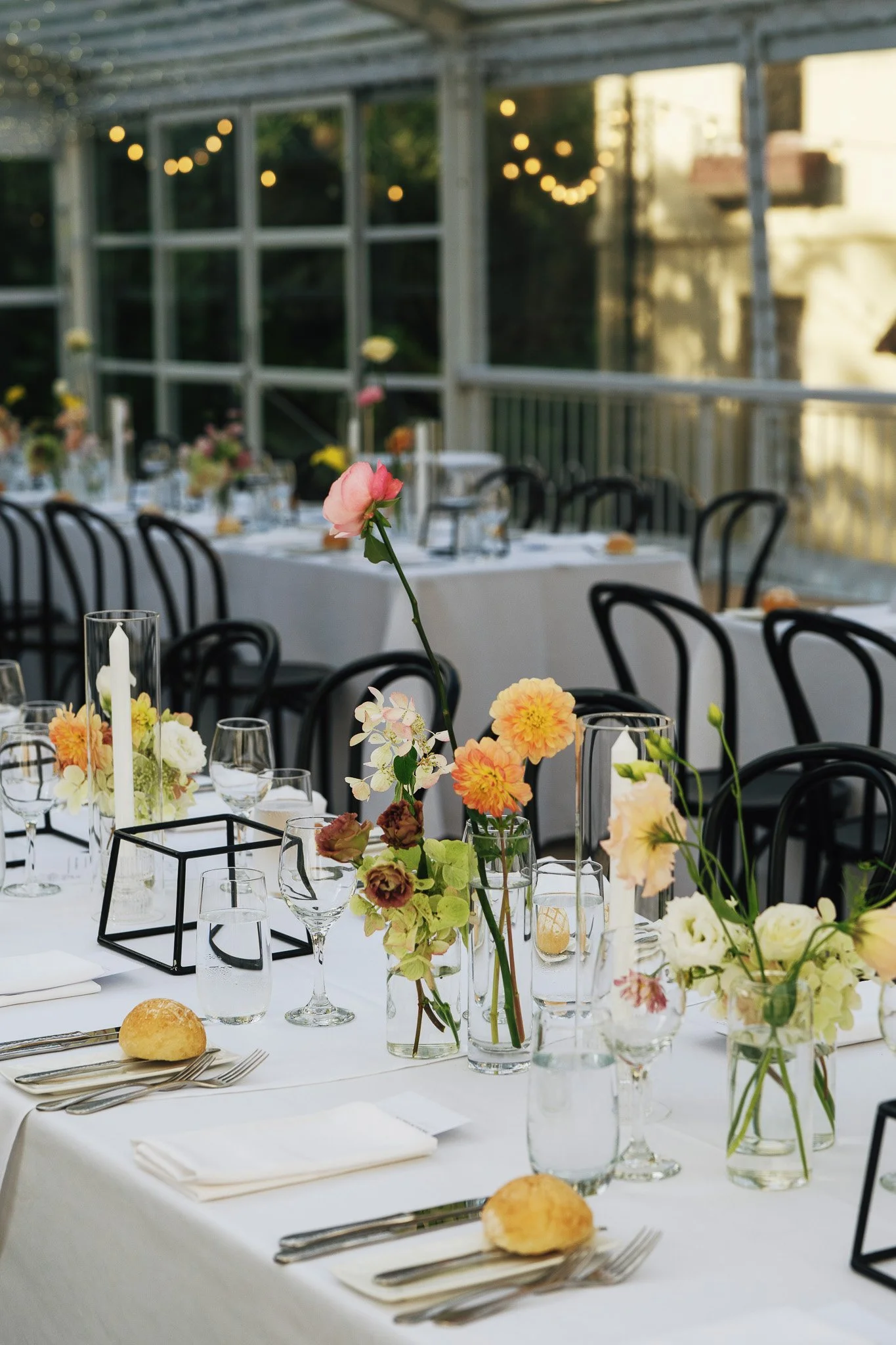 Elegant table setting with flower arrangements, candles, and tableware at a daytime event with string lights inside a glass-walled venue.