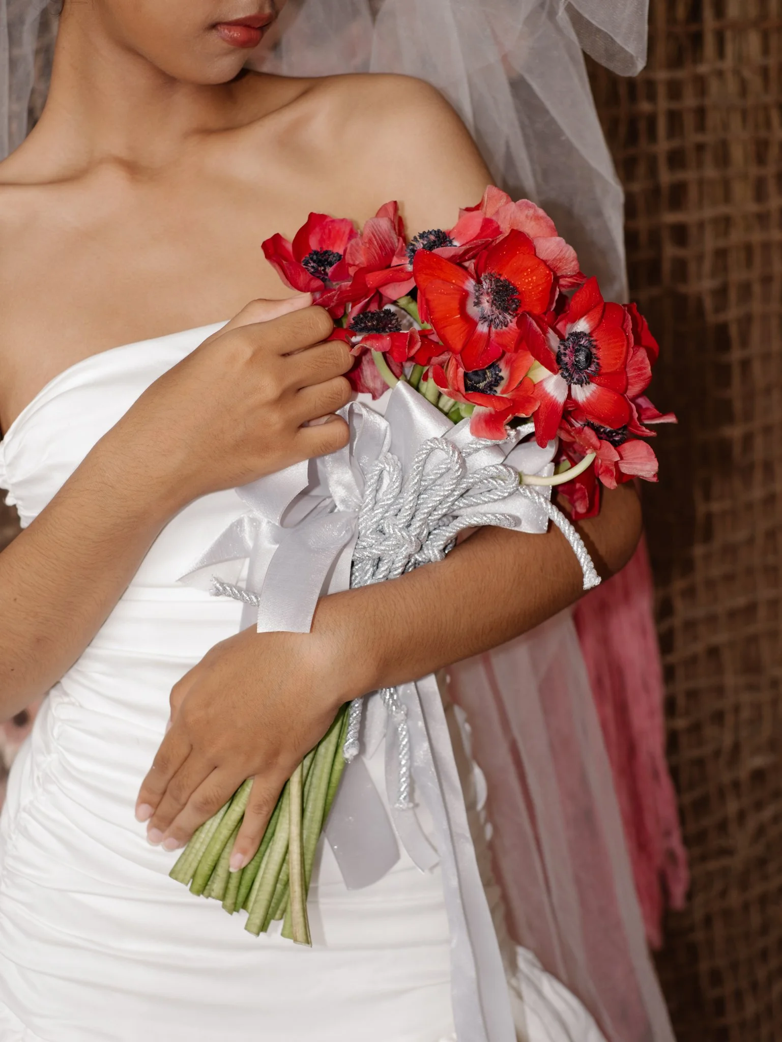 A woman in a wedding dress holding a bouquet of red and pink poppies with black centers. She has her hand on her abdomen and is wearing a veil.