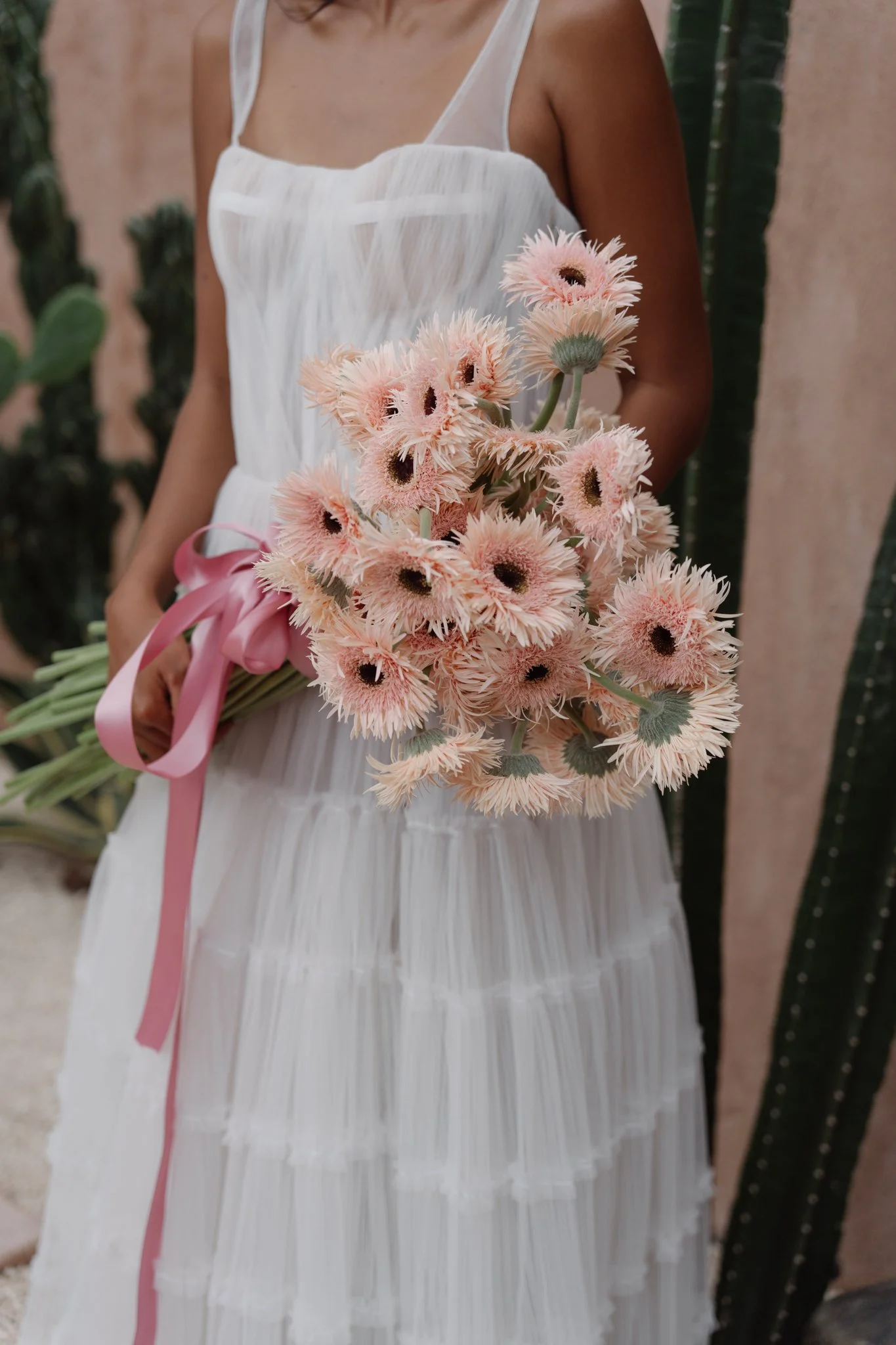 A woman wearing a white dress holding a bouquet of light pink flowers with dark centers, tied with a pink ribbon, standing outdoors with green plants in the background.