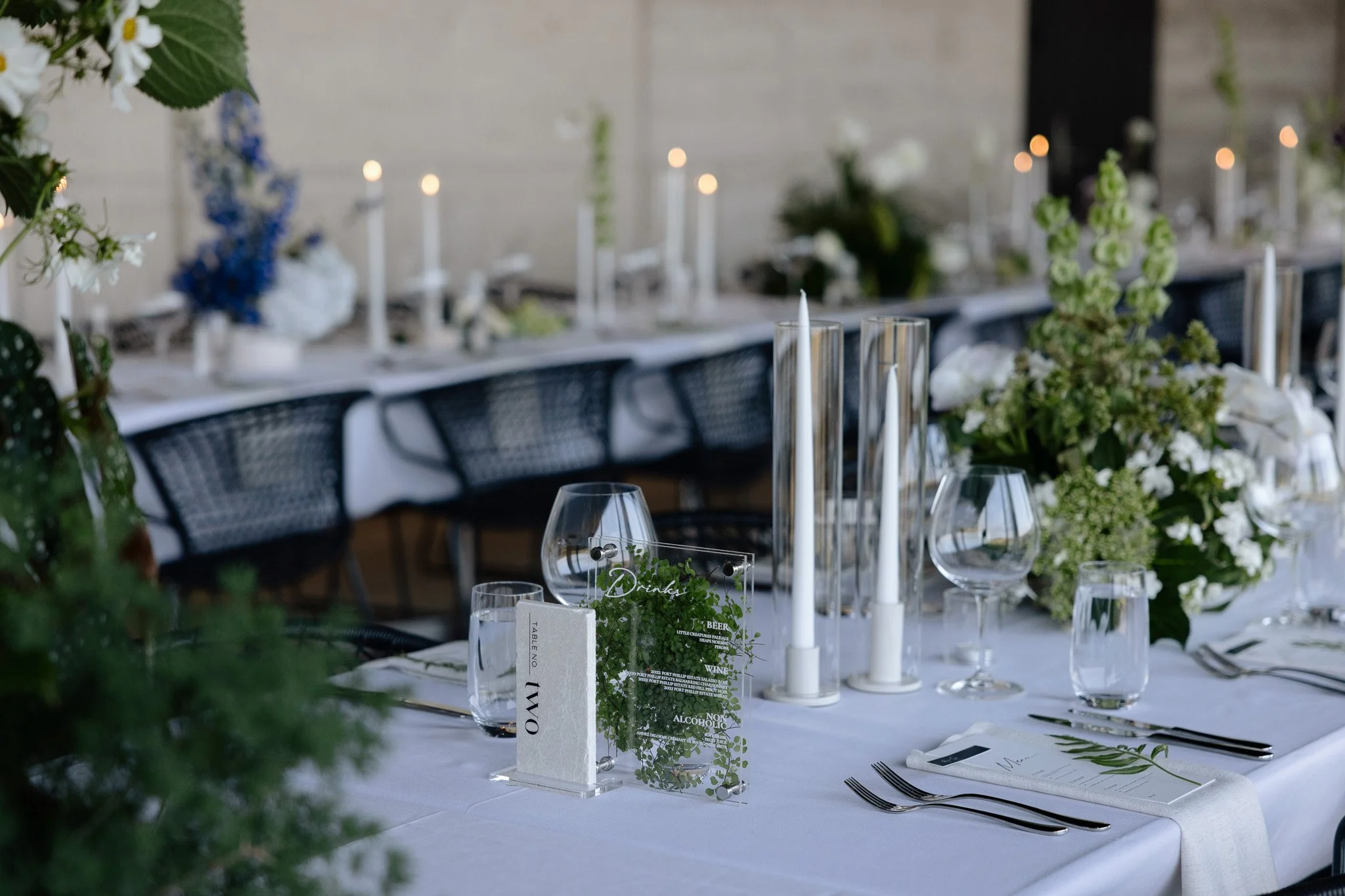 Elegant dining table set with white tablecloth, glassware, silverware, and floral centerpieces in a decorated venue with candles and greenery.