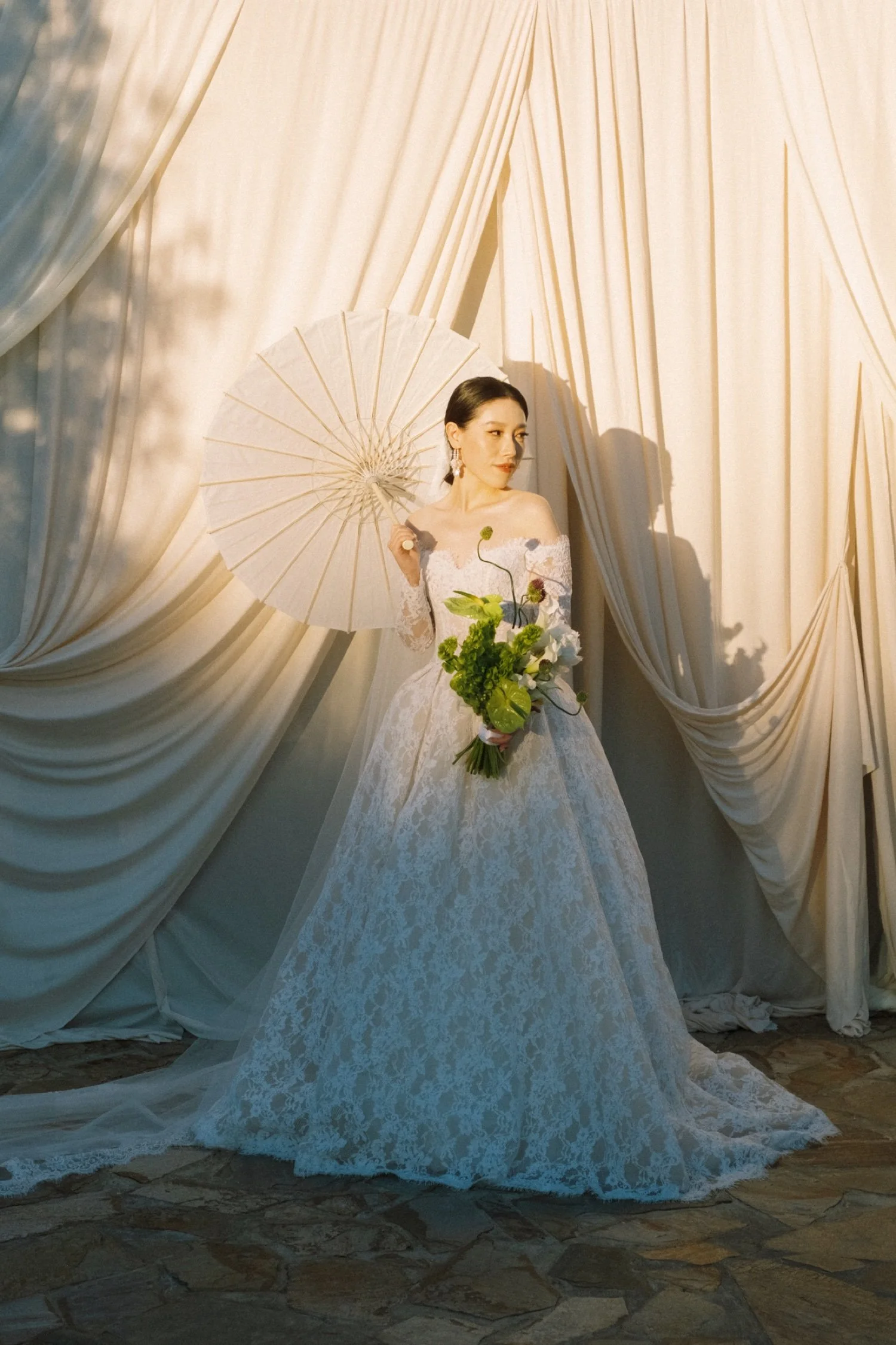 A woman in a white lace wedding dress standing against a backdrop of cream-colored draped curtains. She holds a parasol in her right hand and a bouquet of green and white flowers in her left. The sunlight casts her shadow on the curtains.