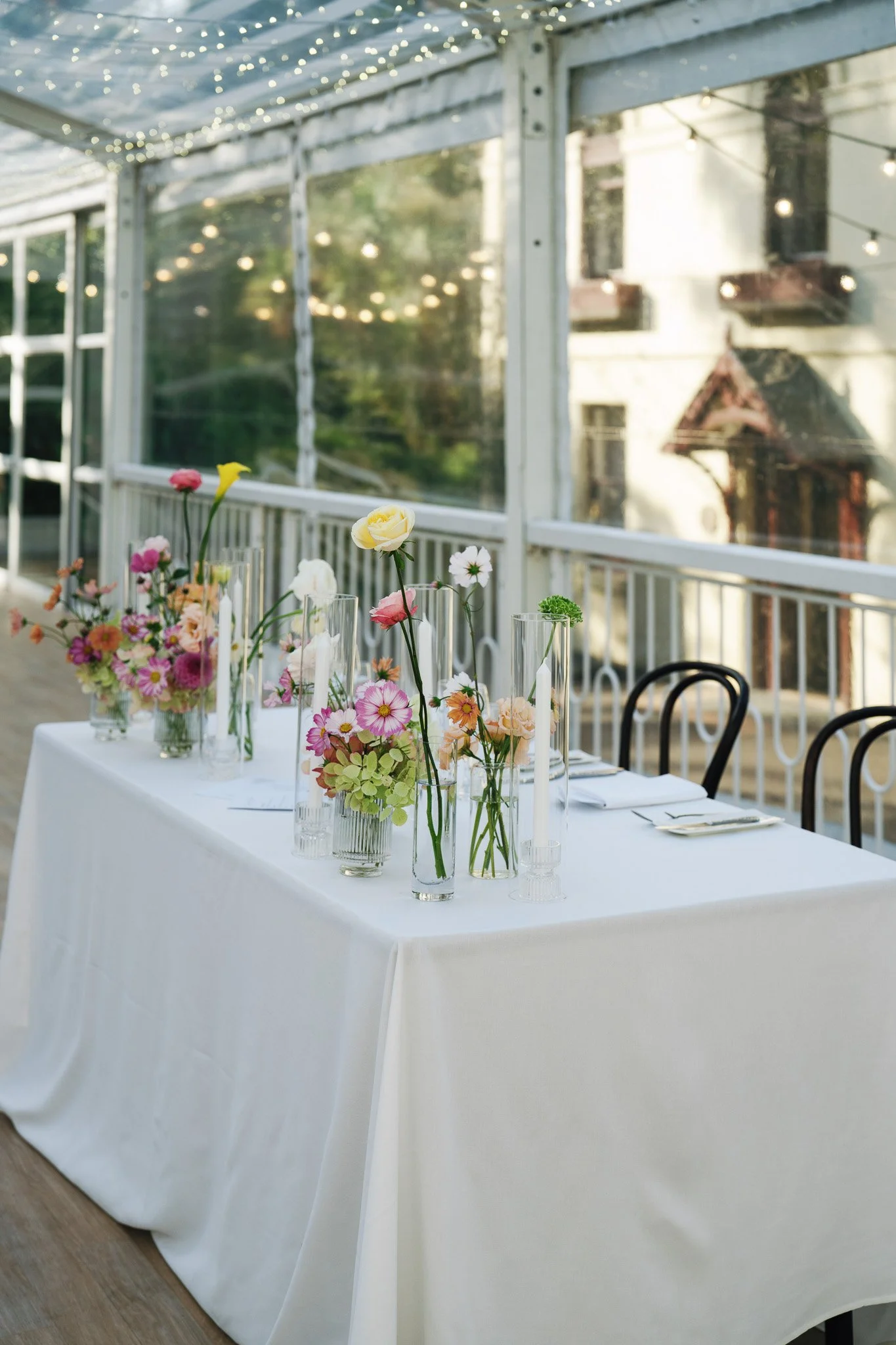 A table decorated with colorful flowers in glass vases and candles, set for a celebration or event, outside under a covered patio with string lights in the background.