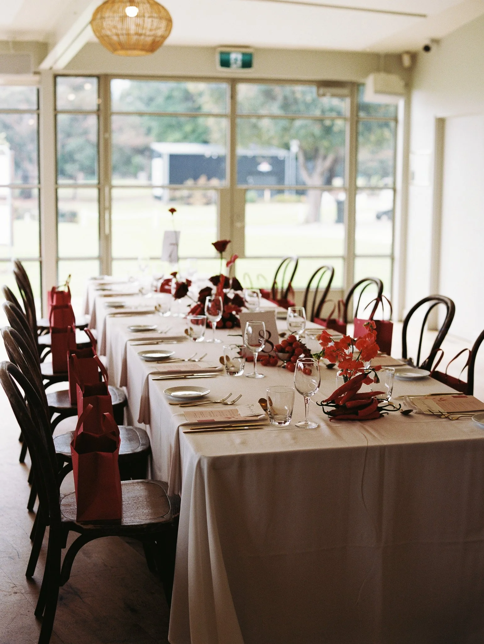 A long dining table with white tablecloths, set with glasses, plates, and utensils, decorated with red flowers, in a room with large windows revealing an outdoor view, and red gift bags on chairs.