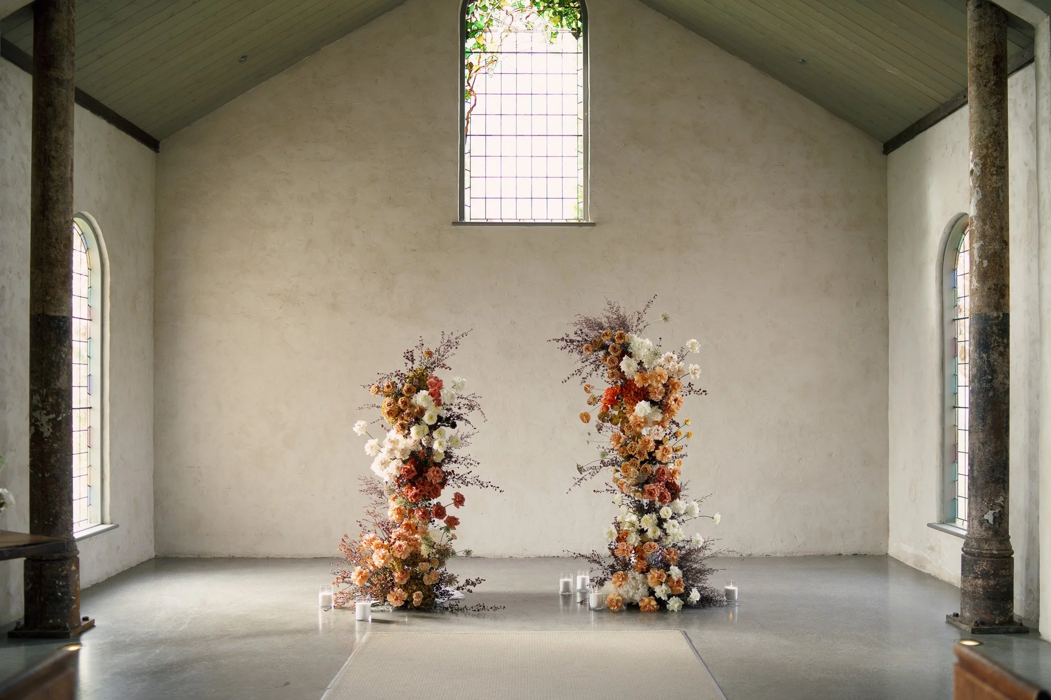 Interior of a church with a high ceiling and stained glass windows. Two tall floral arrangements with orange, white, and peach flowers are in front of the altar, surrounded by small white candles.