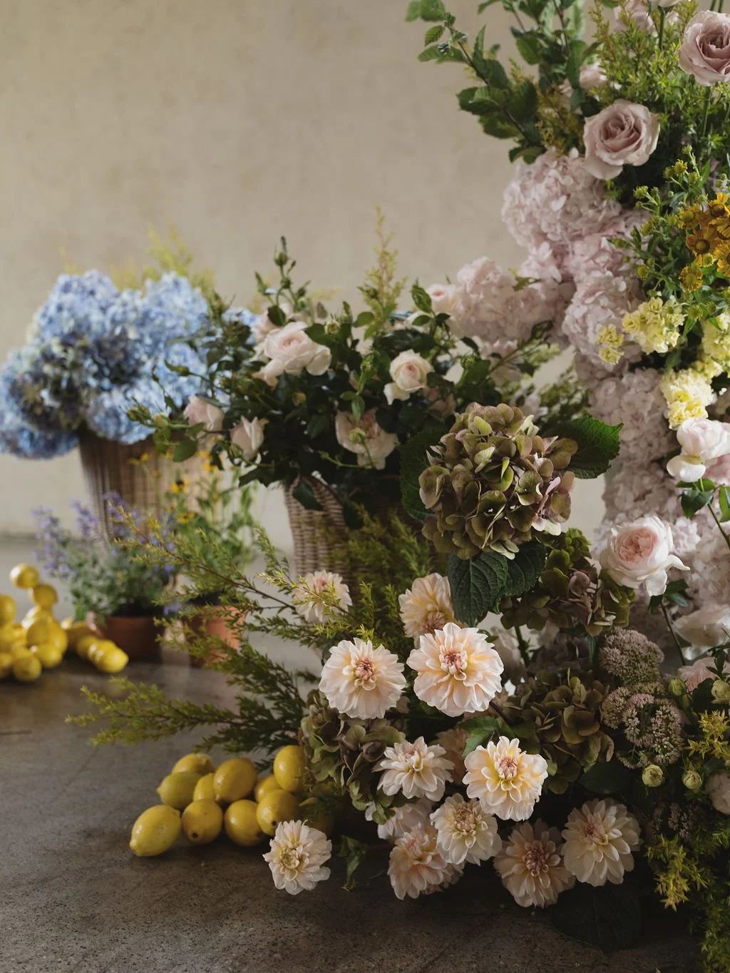 Arrangement of pastel-colored flowers including pink roses, blue hydrangeas, white dahlias, and yellow accents in baskets and on a table with yellow lemons.