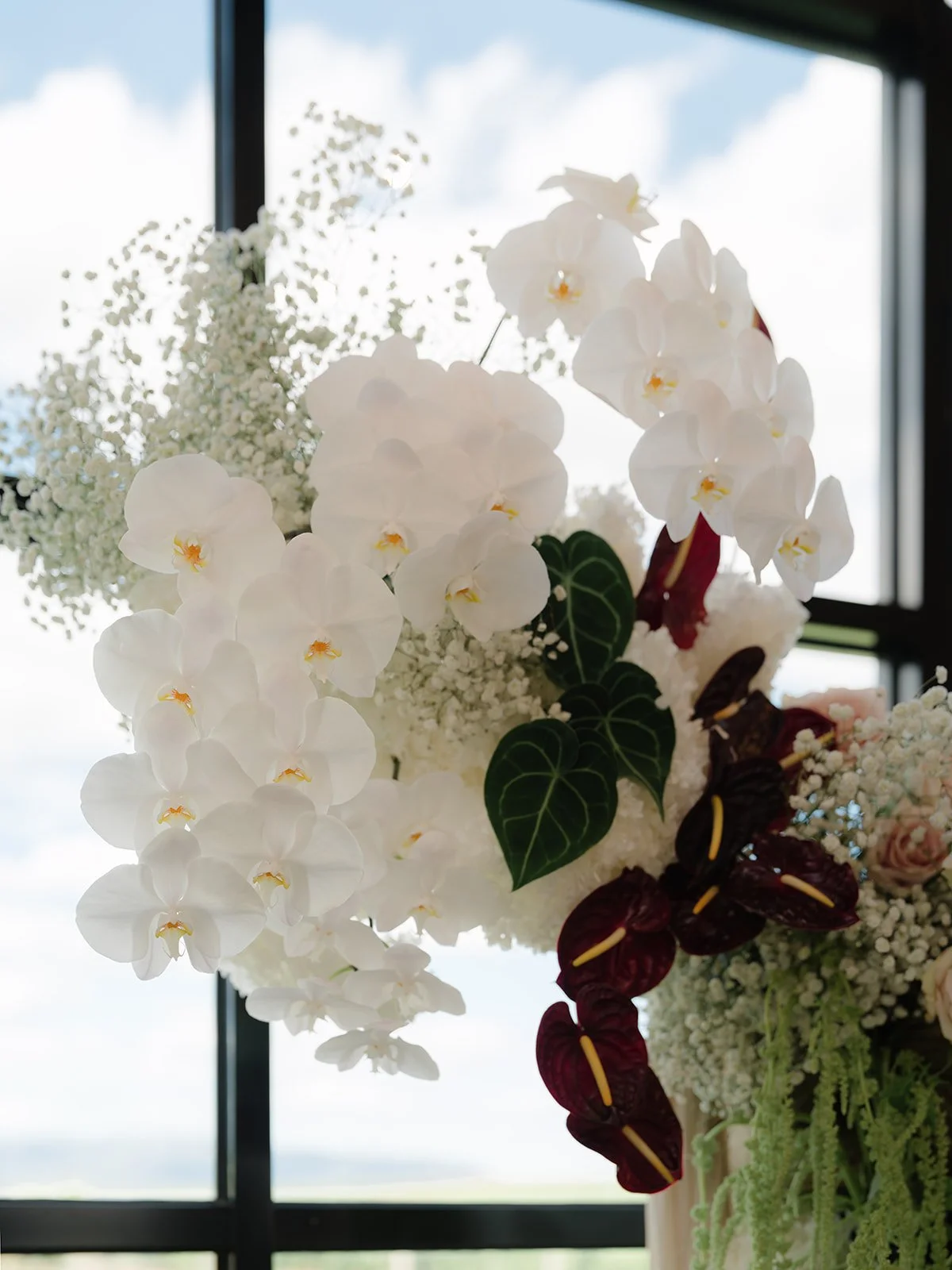 Close-up of a floral arrangement with white orchids, dark red anthuriums, green leafy plants, and other white flowers against a window with a partly cloudy sky.
