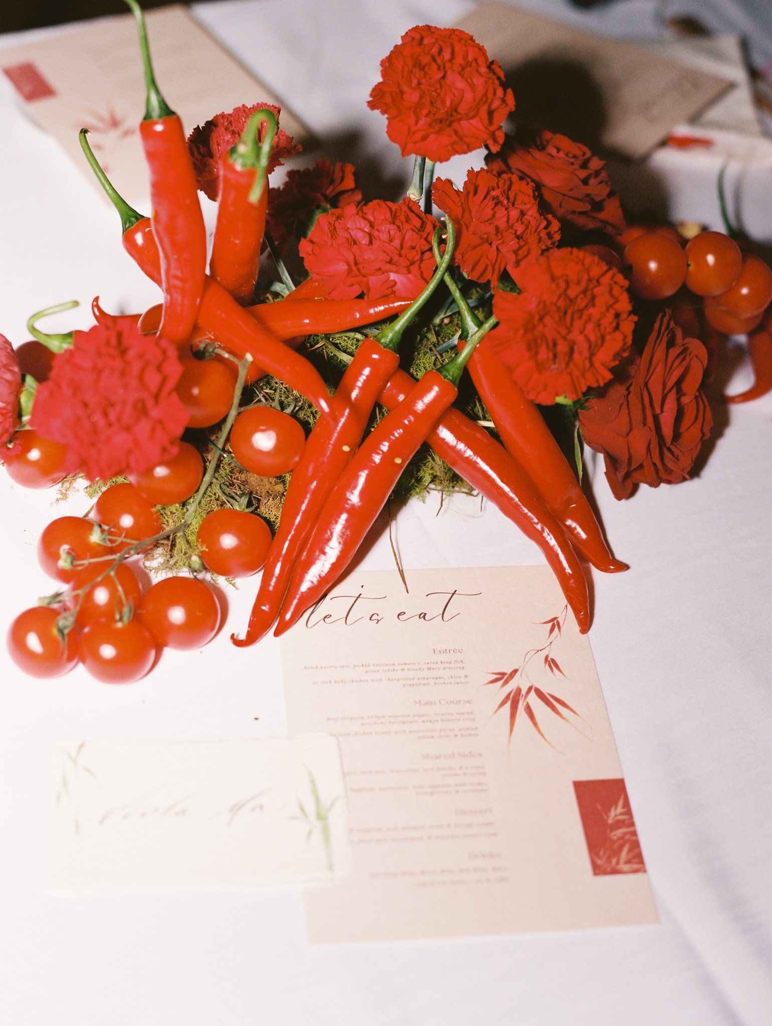 A close-up of a decorative arrangement of red chili peppers, red cherry tomatoes, and red carnation flowers on a restaurant menu or paper, with a white background.
