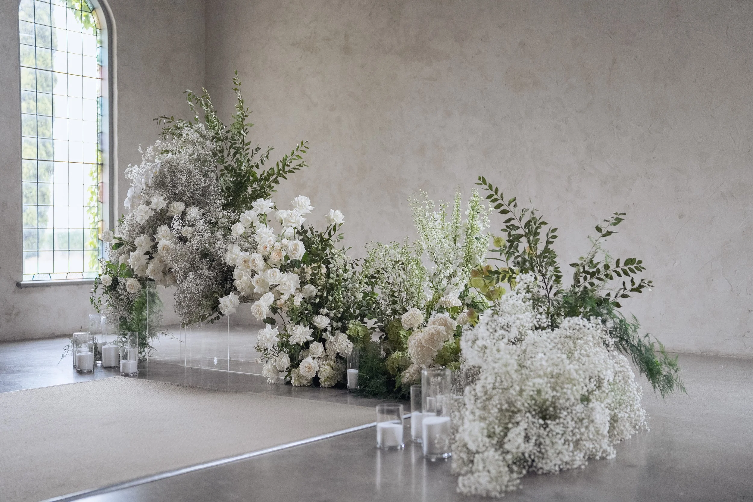Large floral arrangement with white and green flowers and foliage, placed on a white carpeted floor in a room with a large arched window and plain wall, surrounded by glass candle holders.