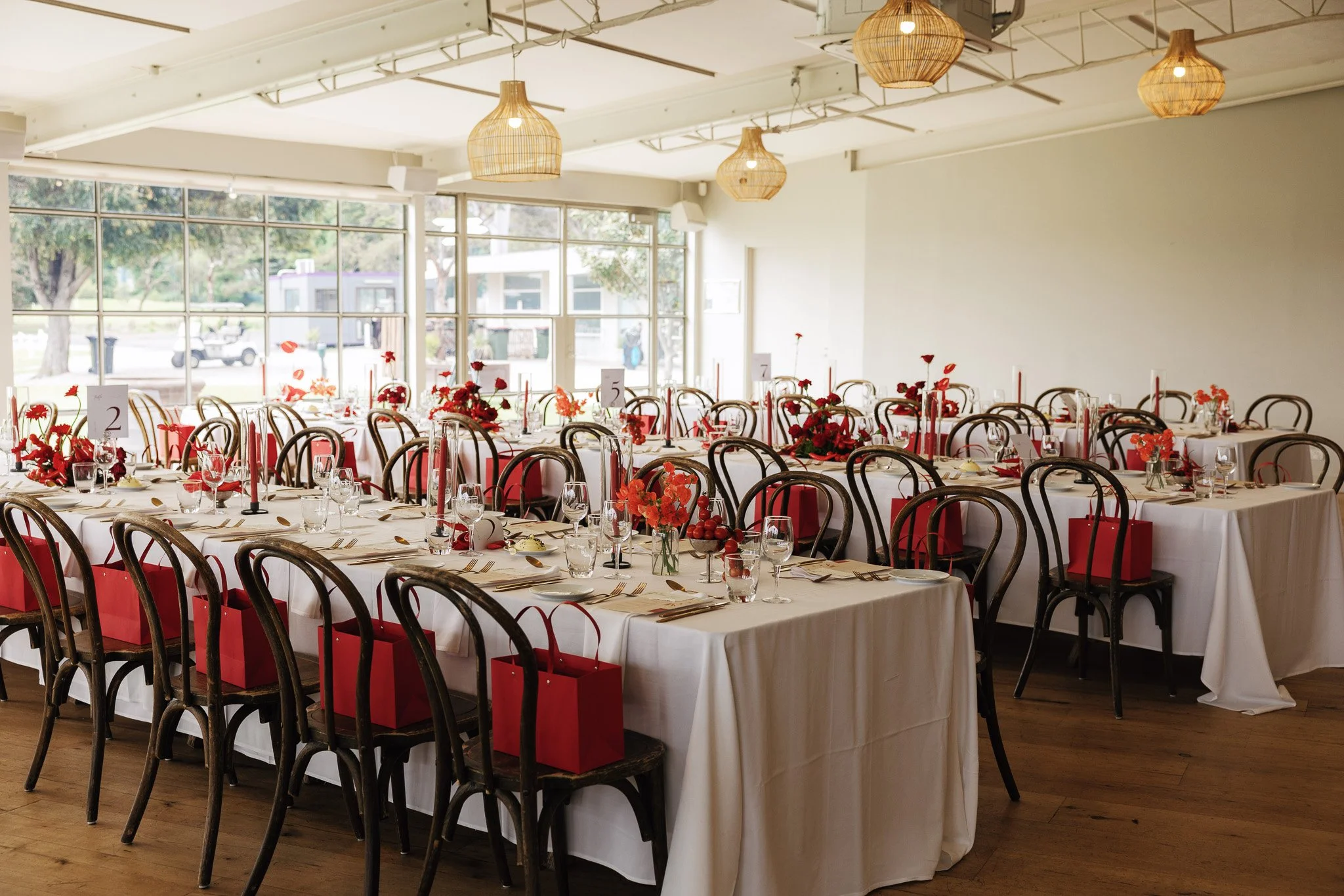 A large indoor banquet hall set up for a wedding or special event with long tables decorated with red floral centerpieces, candles, and tableware. The hall has large windows, wooden chairs, and hanging woven light fixtures.