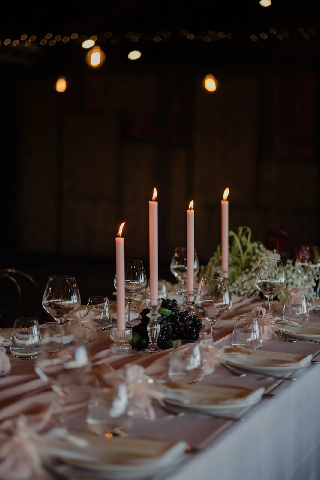 Elegant dinner table decorated with pink candles, wine glasses, white plates, and a floral centerpiece with grapes, illuminated by soft candlelight in a dark room.