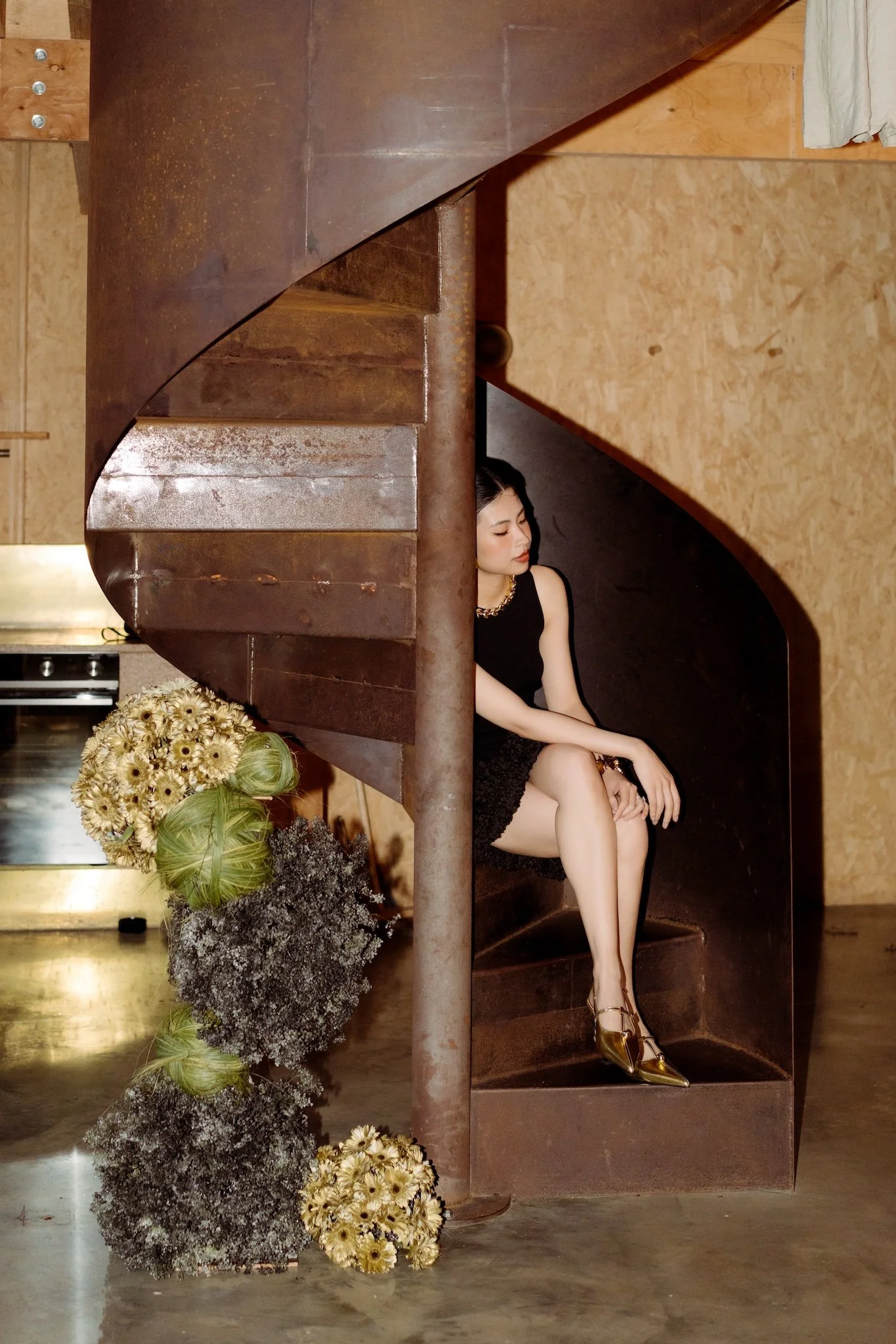 A woman in a black dress sitting on a wooden staircase, surrounded by flower arrangements, with a rustic indoor setting.
