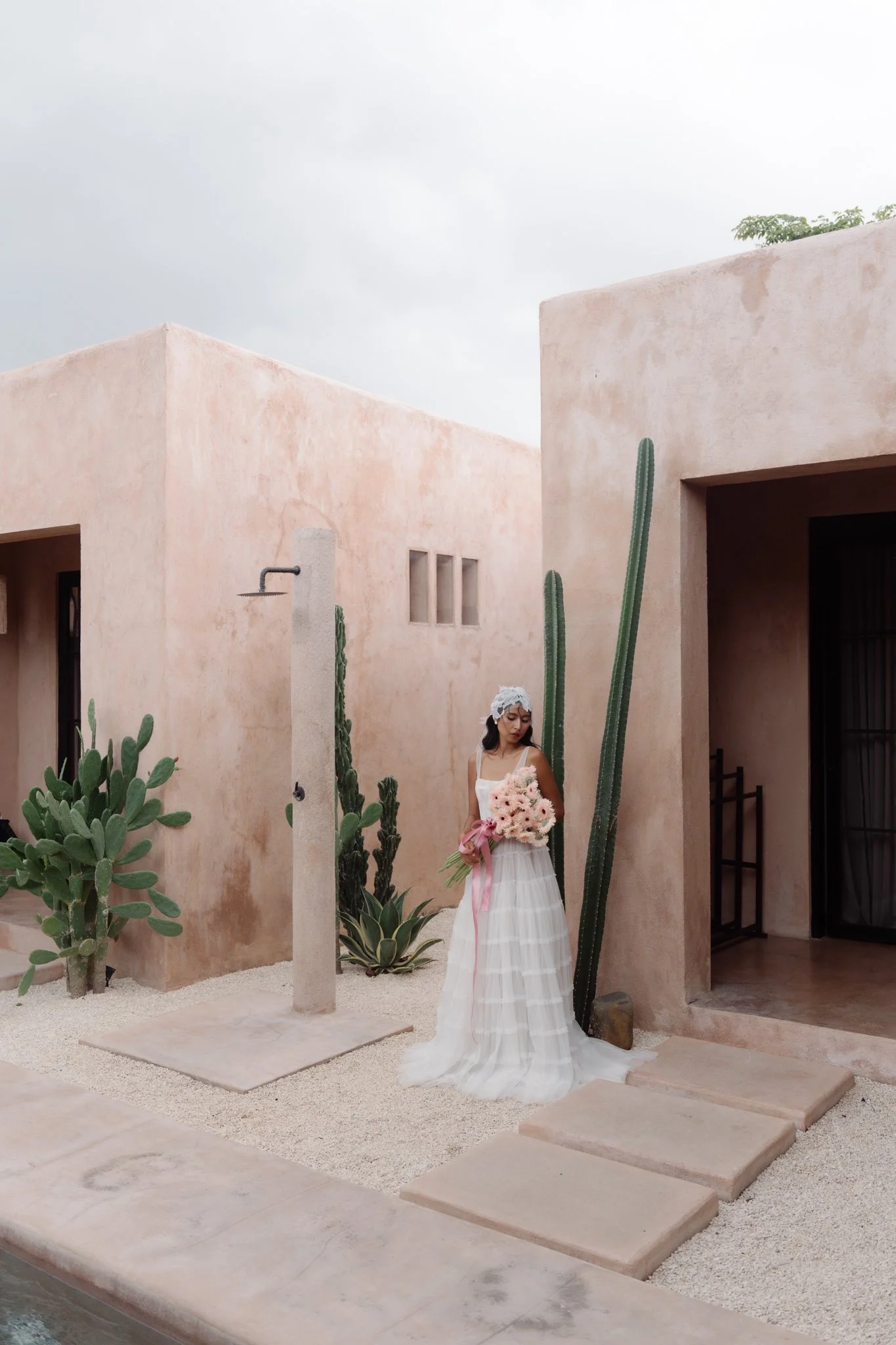 A bride in a white wedding dress holding a large bouquet, standing on a stone step outside a peach-colored building with desert plant landscaping and a drill shower nearby.