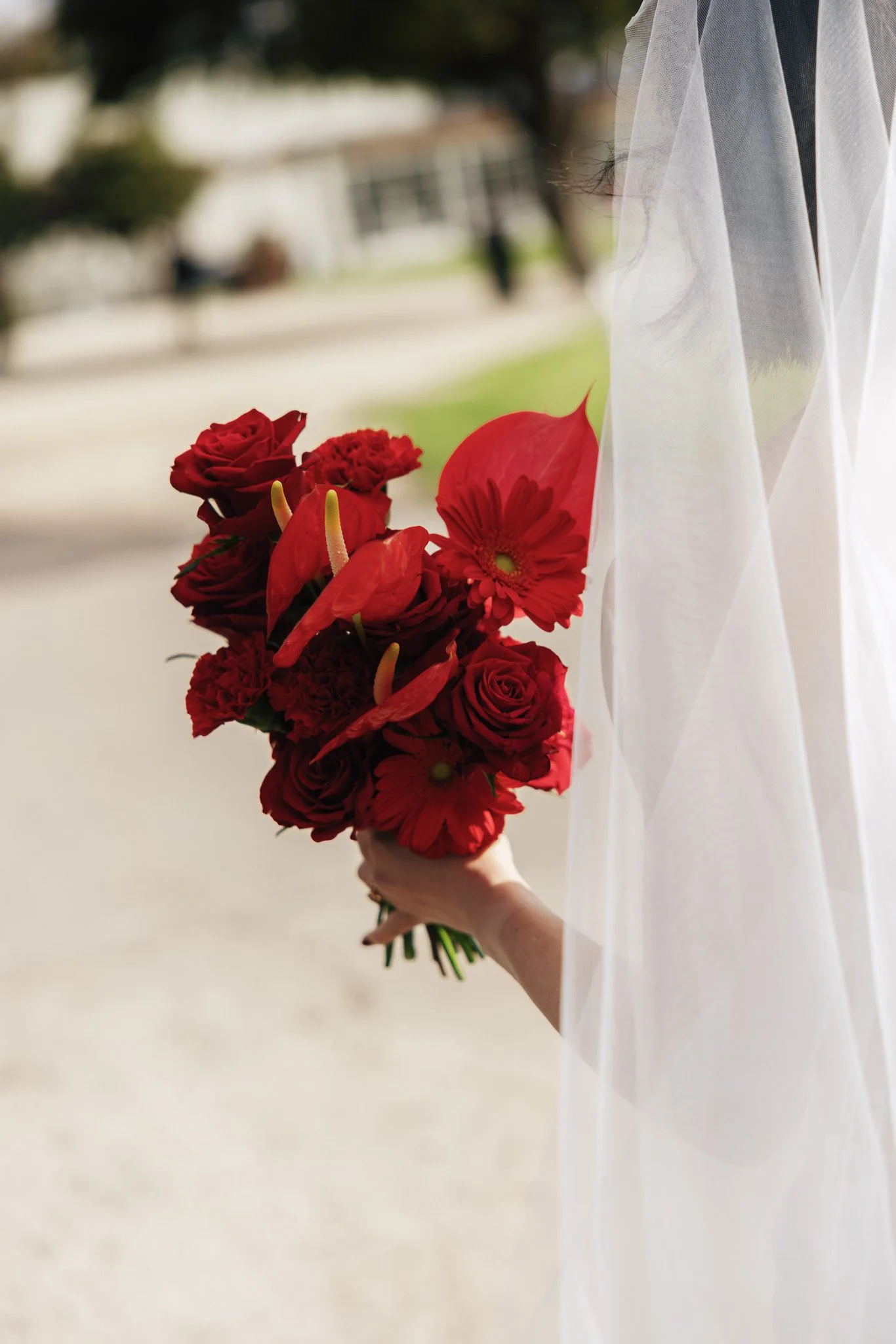 A person in a white dress holds a bouquet of red flowers, standing outdoors near a window with sheer white curtains.