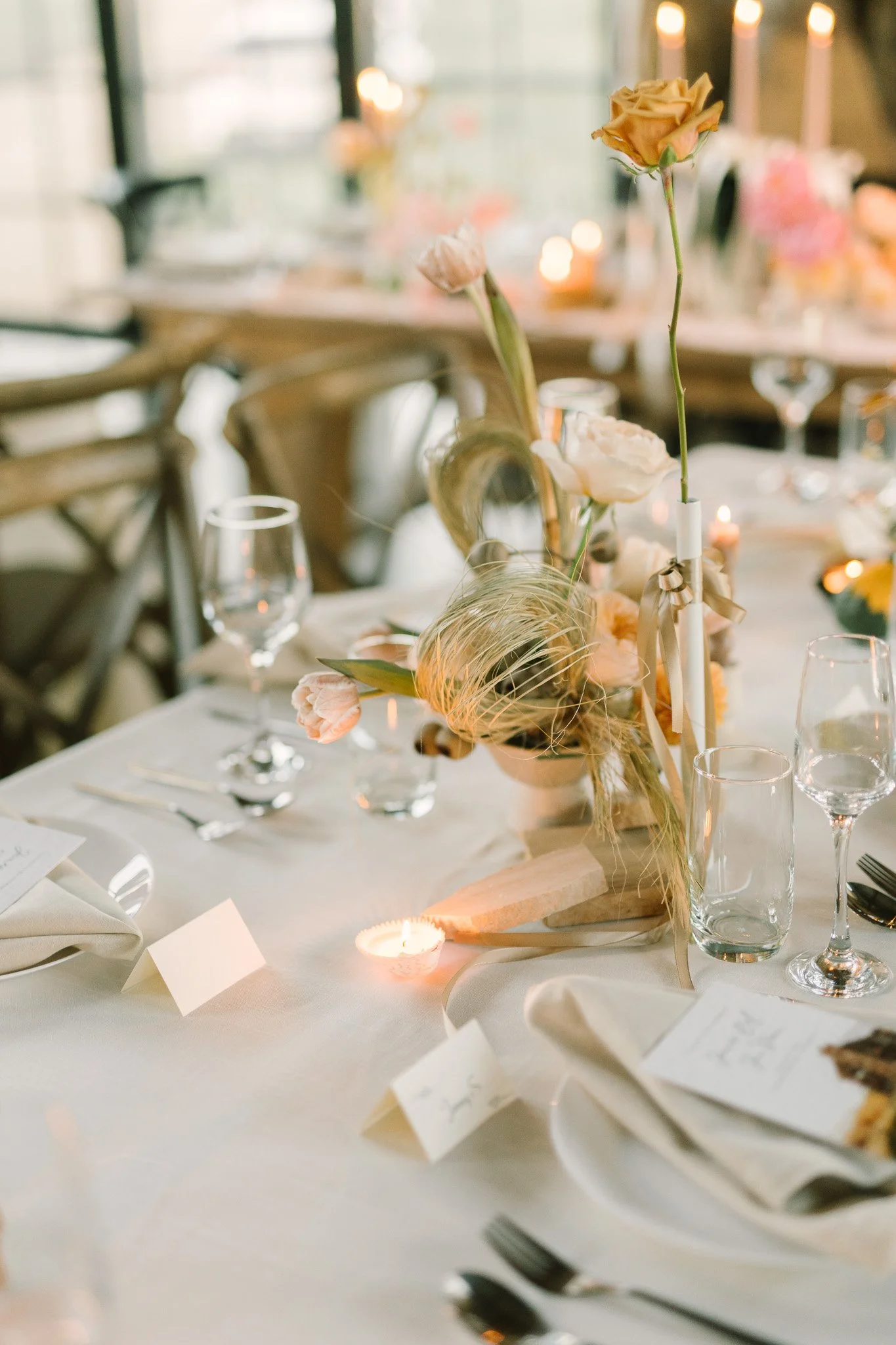 A decorated table for a wedding or special event featuring a floral centerpiece with roses and other flowers, surrounded by wine glasses, candles, and place cards on a white tablecloth, with chairs in the background.