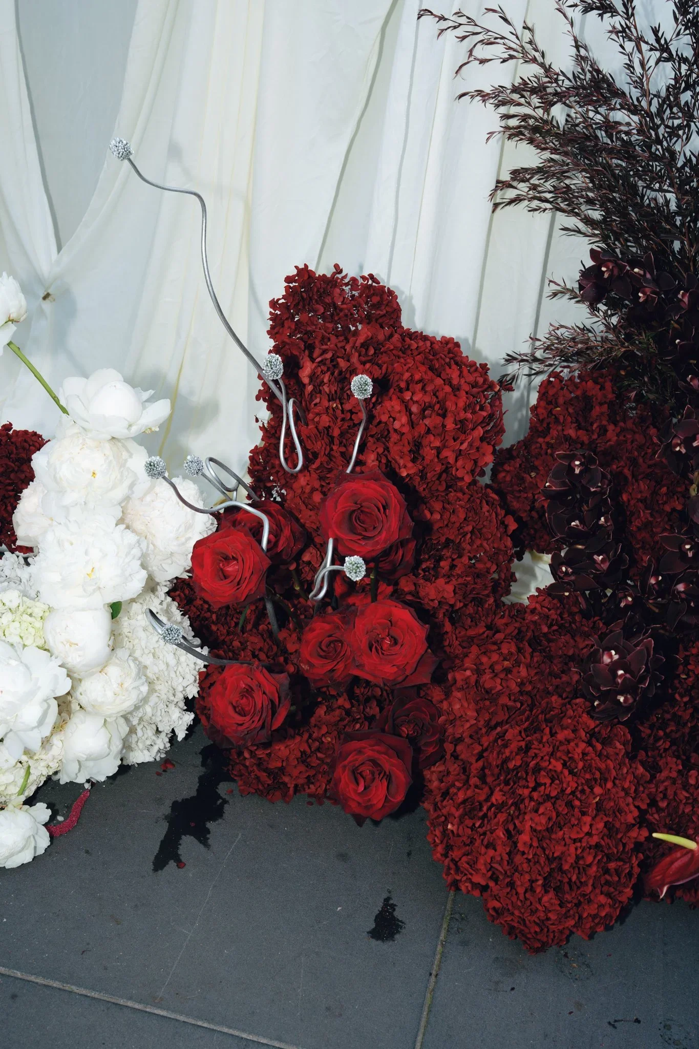 A display of various flower arrangements, including white, red, and dark purple flowers, against a light-colored curtain backdrop.