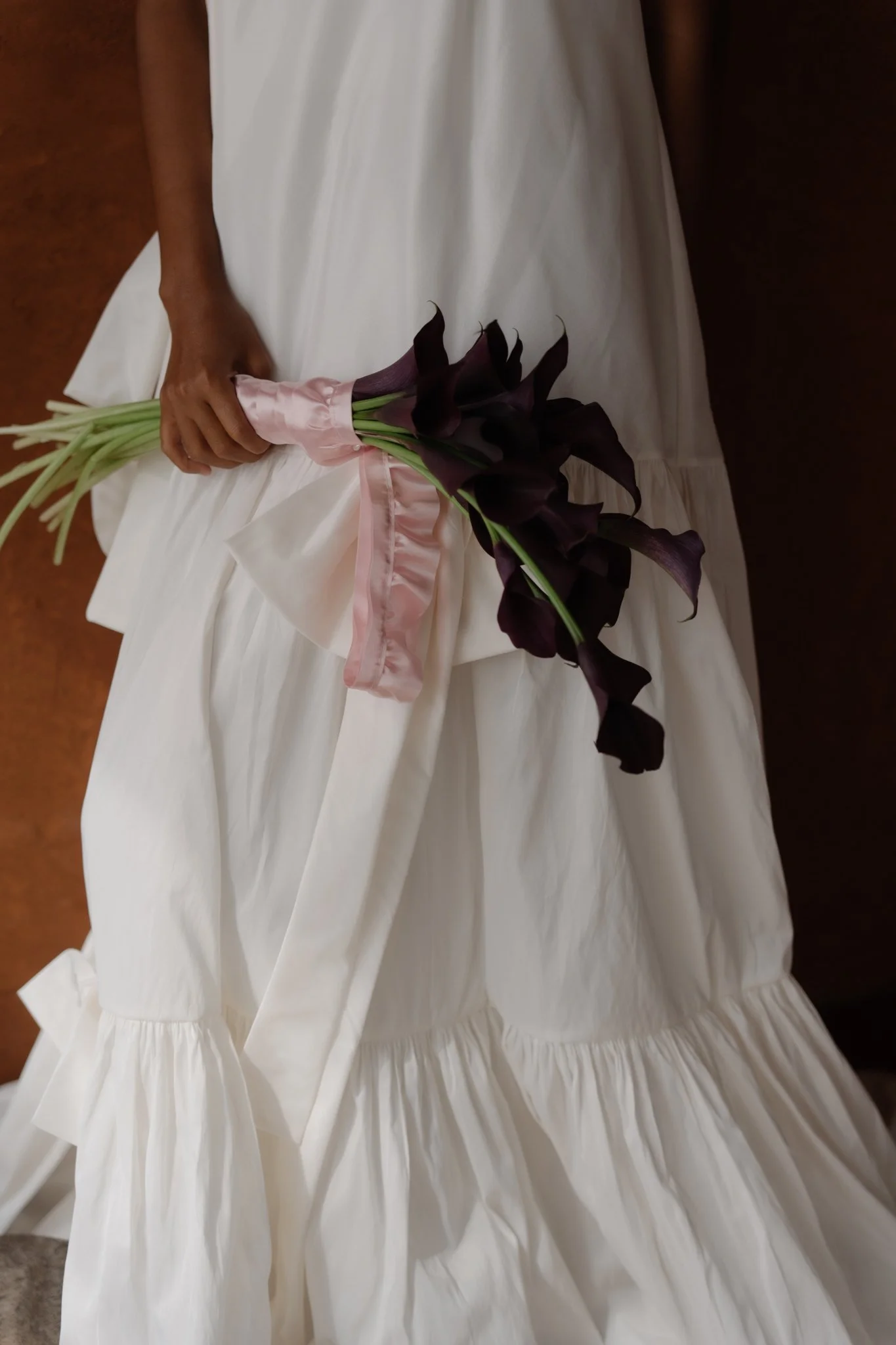 A person in a white dress holding a bouquet of dark purple calla lilies wrapped in pink satin ribbon.