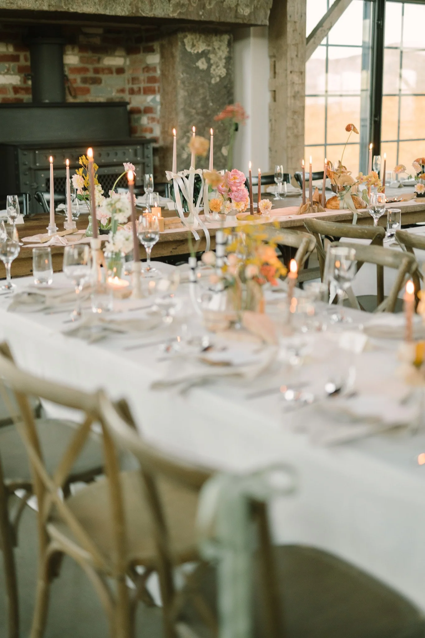 A long dinner table decorated with flowers, candles, and tableware inside a rustic brick and wood building with large windows.