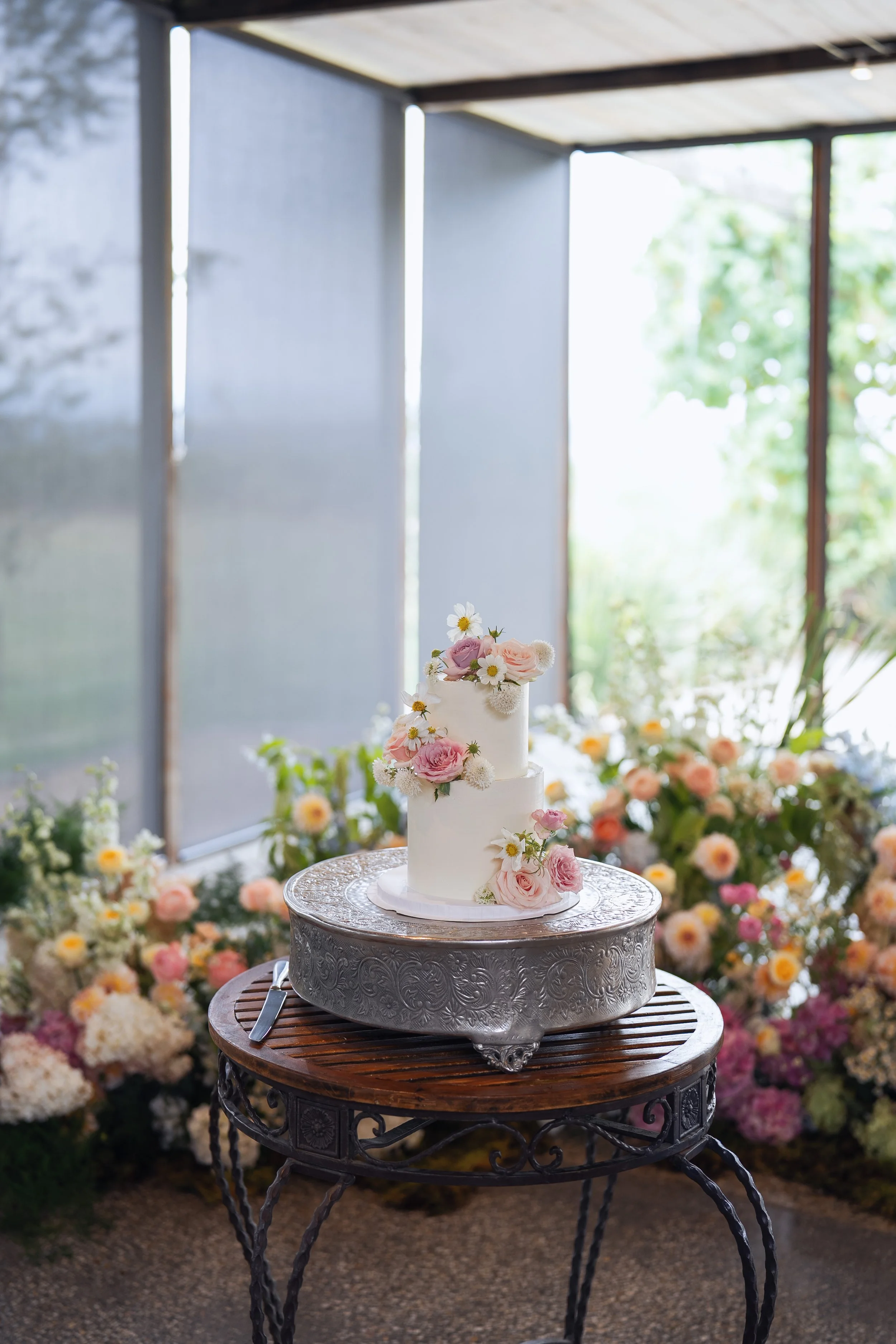 A three-tiered white wedding cake decorated with pink and white roses and daisies, set on a silver ornate cake stand on a round wooden table, surrounded by colorful flowers and greenery, in a bright, natural light setting.