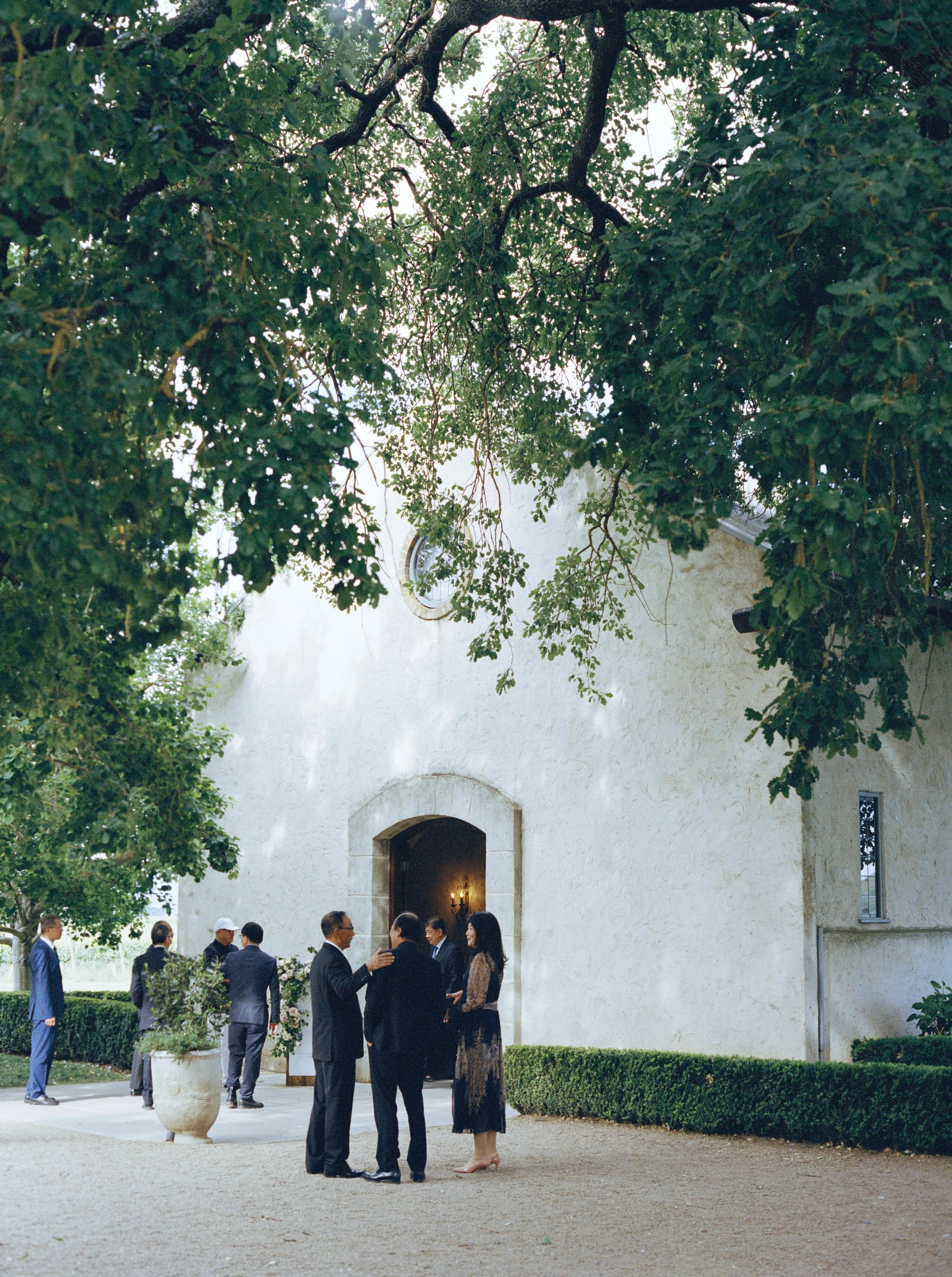 A group of people dressed in formal attire socializing outside a white stucco building with an arched entryway, surrounded by greenery and trees.
