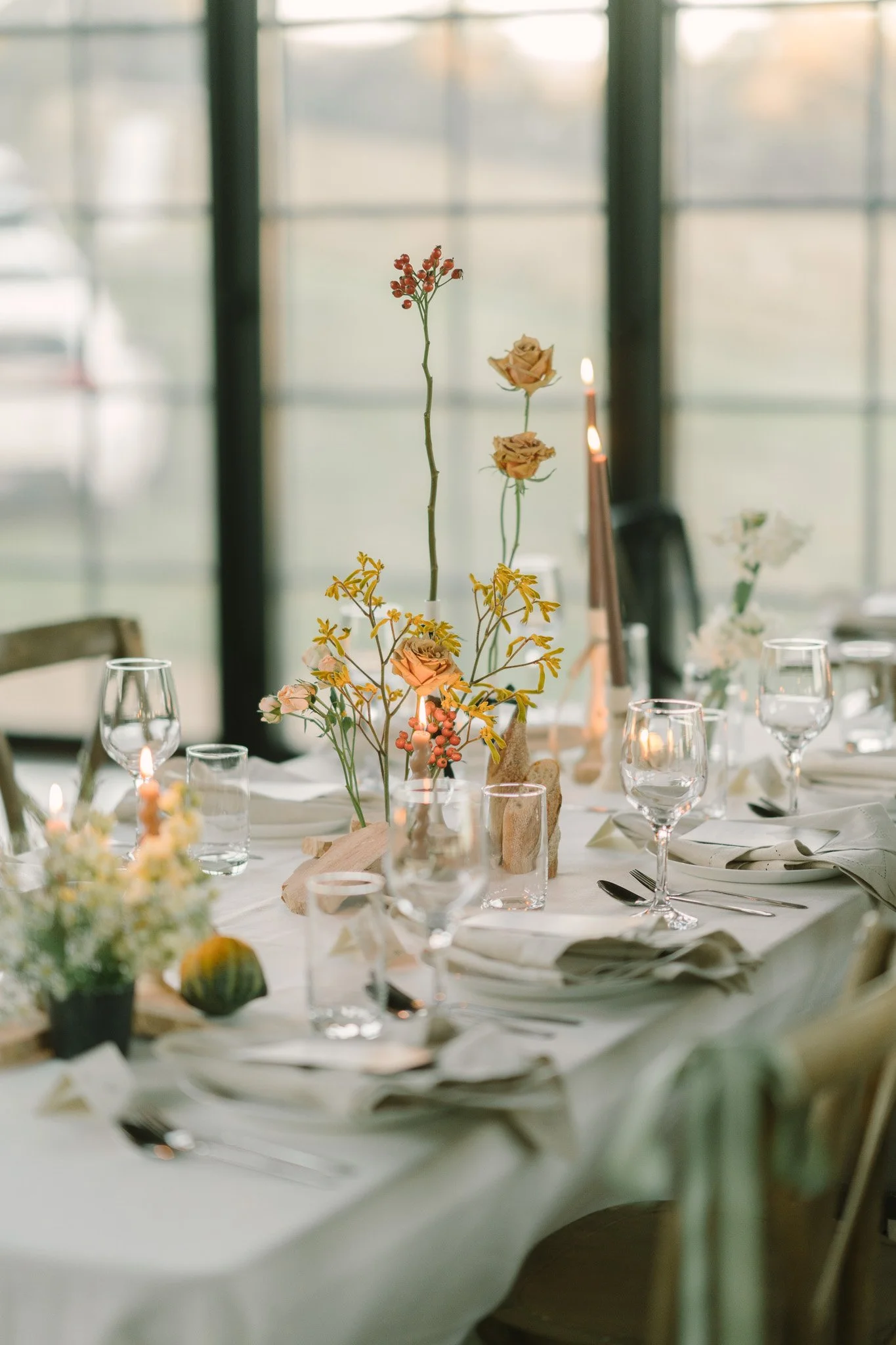 A decorated dining table with floral centerpieces, wine glasses, plates, and cutlery, set in a softly lit room with large windows in the background.