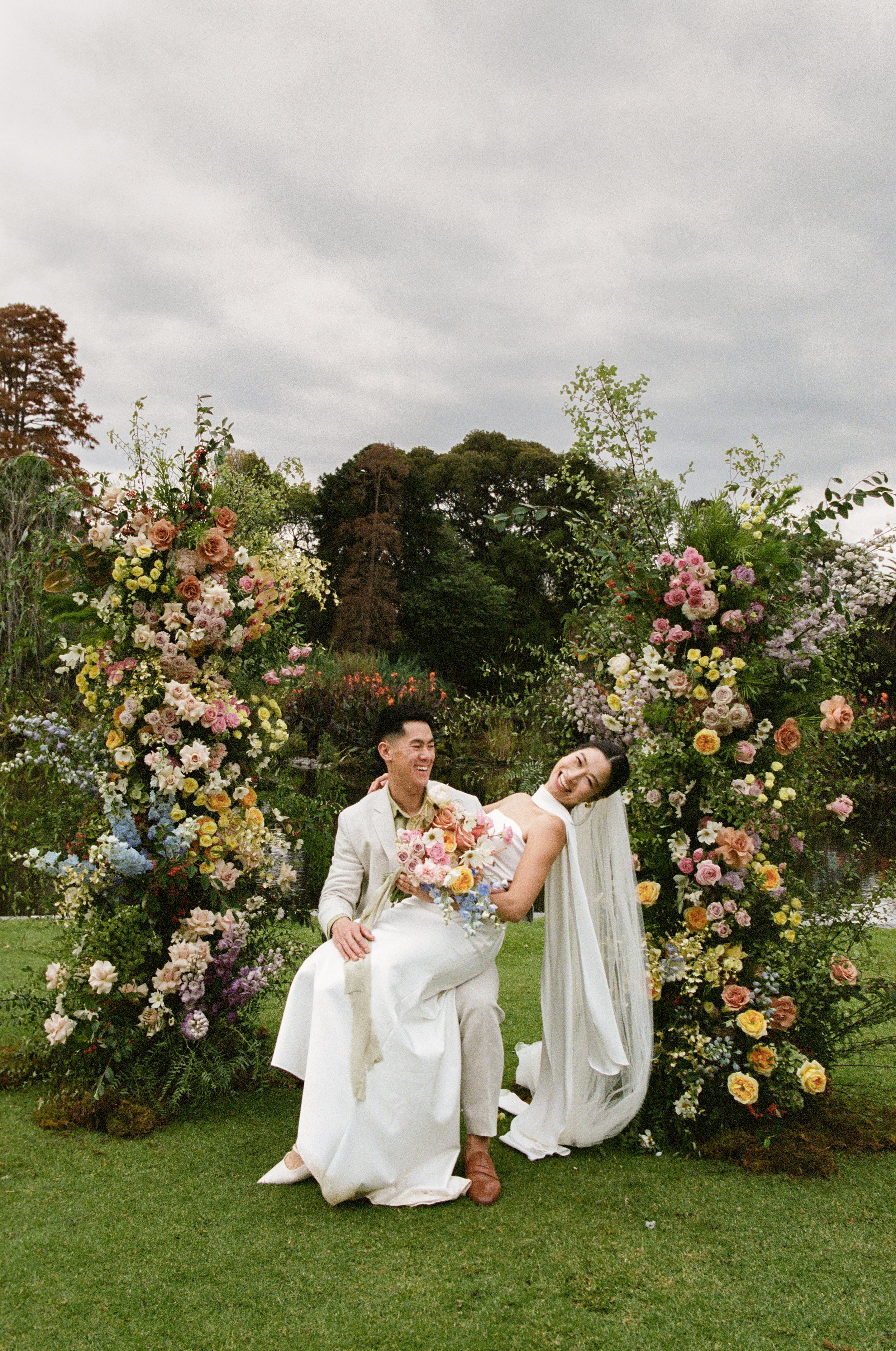 A happy couple in wedding attire, smiling and sitting close together, surrounded by an arch decorated with colorful flowers outdoors on a cloudy day.
