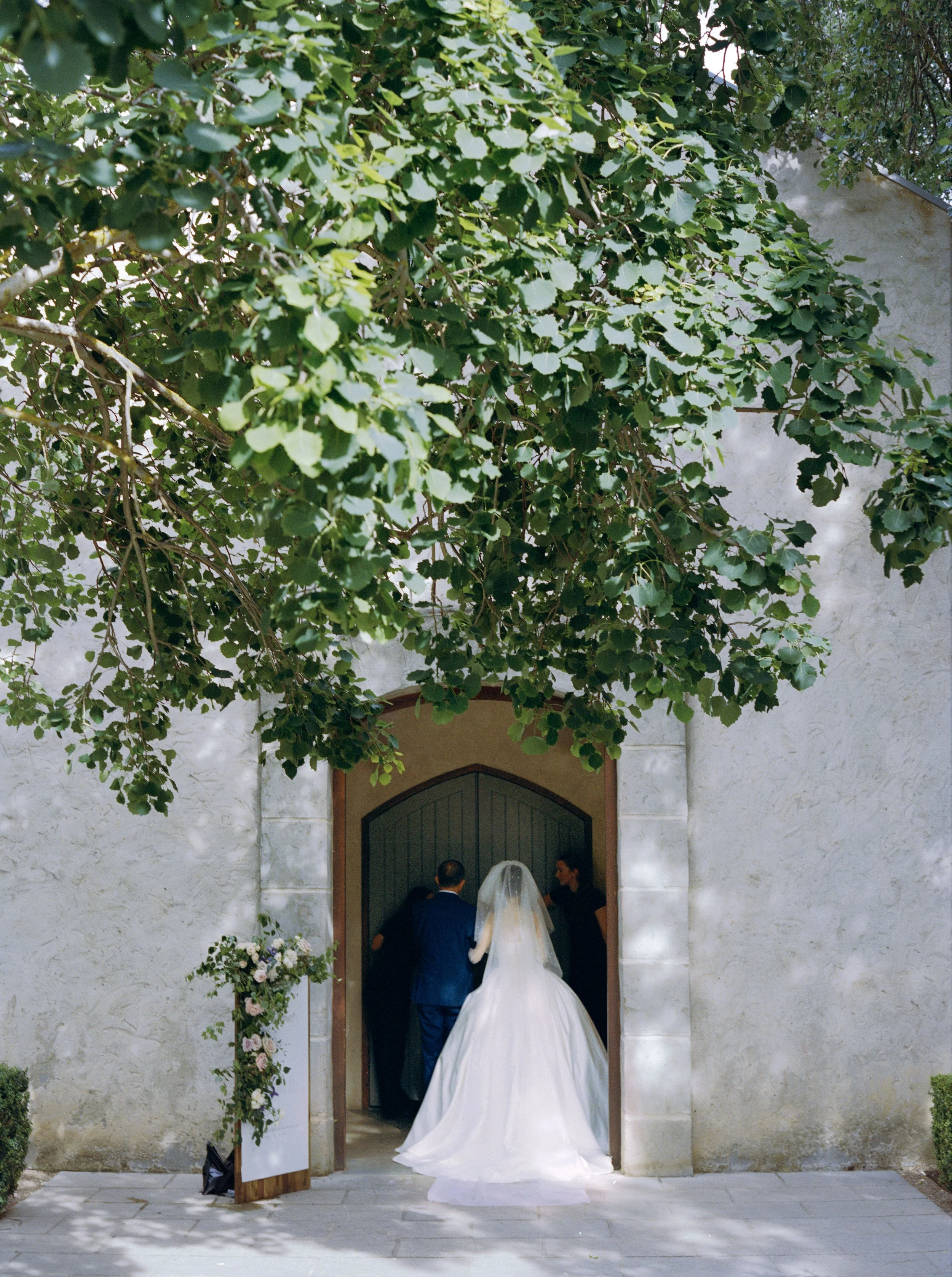 A bride and groom entering a wedding venue through an arched doorway, with a wedding officiant and a woman on the right side. The scene is framed by green leafy branches overhead and a white wall with a stone texture.